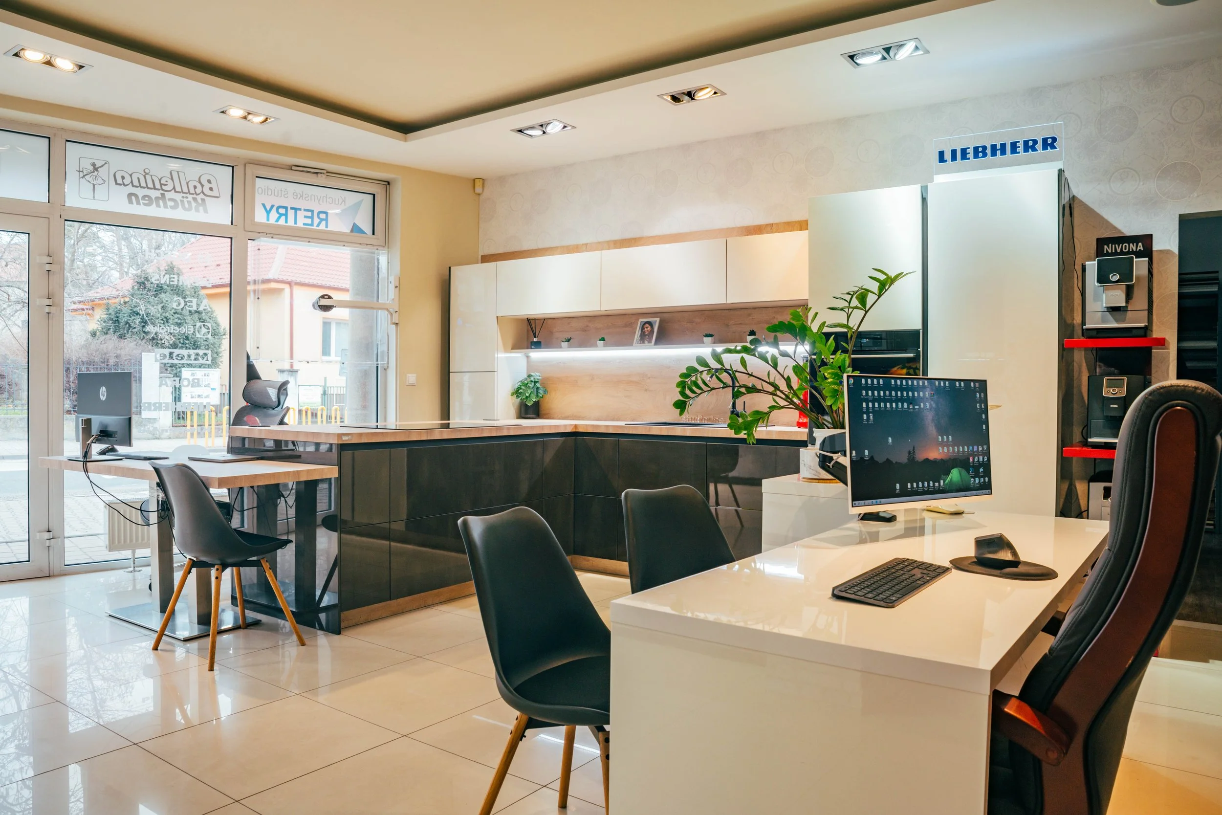 Office space with desks, chairs, computers, and large windows, illuminated by ceiling lights and natural light from the windows.