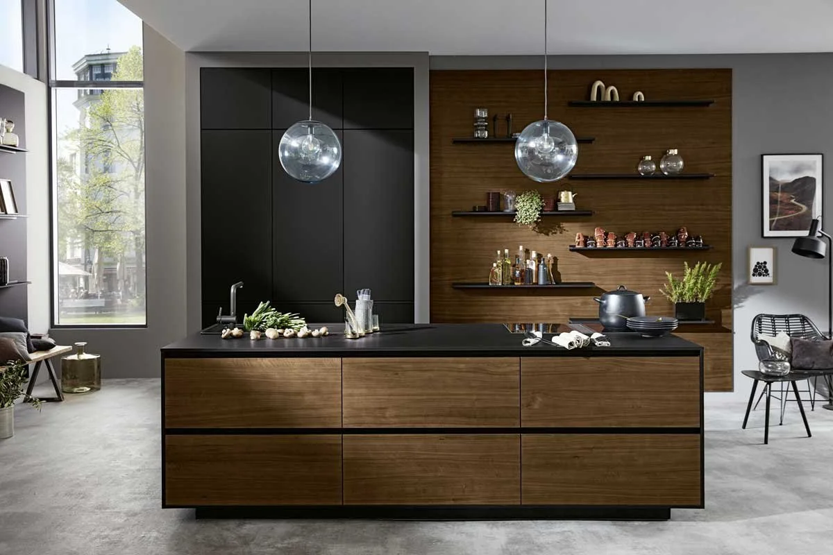 Modern kitchen with black and wood cabinetry, open shelves displaying bottles and decor, two hanging glass pendant lights, a large window with trees outside, and a small seating area on the right.