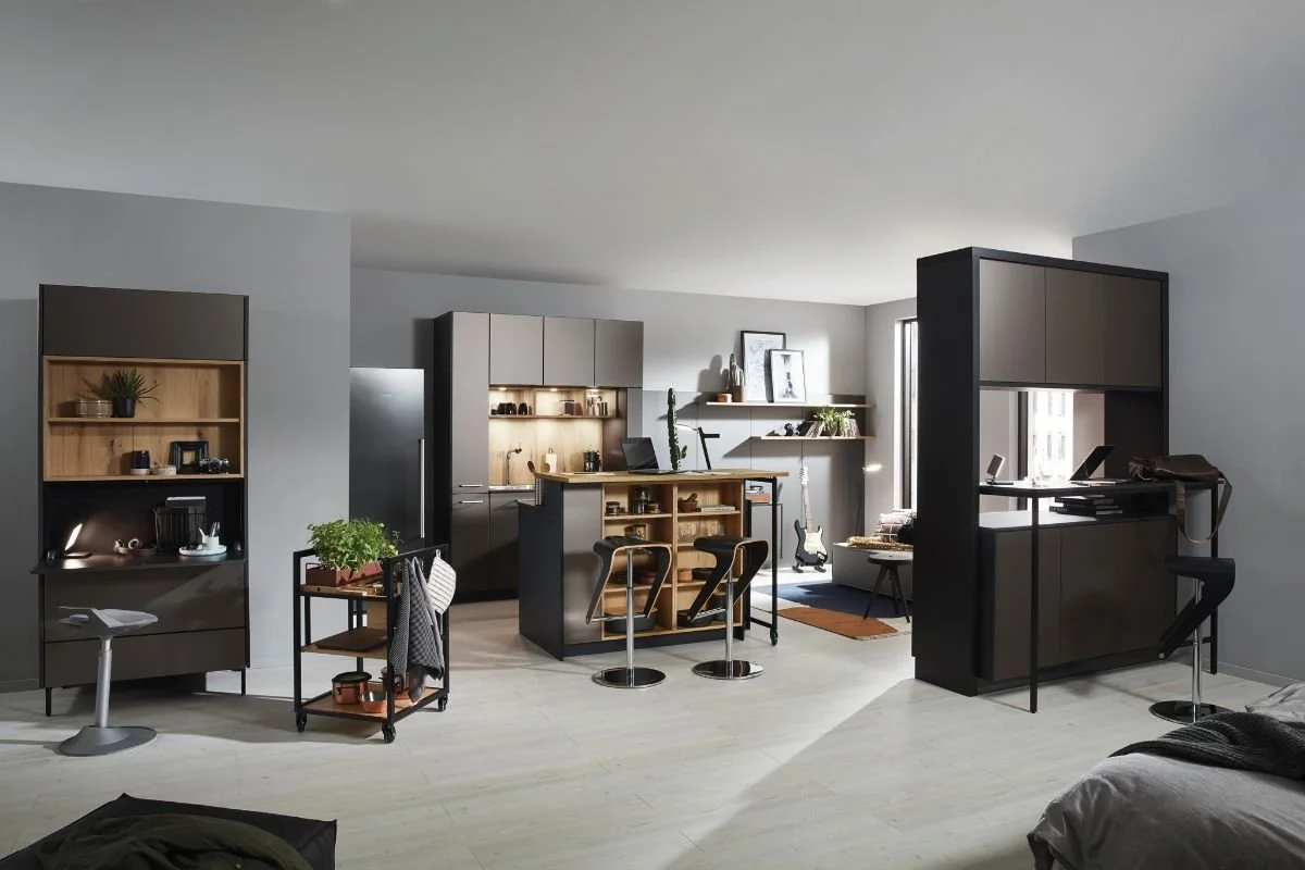Modern open-concept living space with kitchen, dining, and sitting area. The kitchen has black and beige cabinets, a central island, and shelves with decor and plants. Furniture includes bar stools, a small table with plants, and a lounging area with