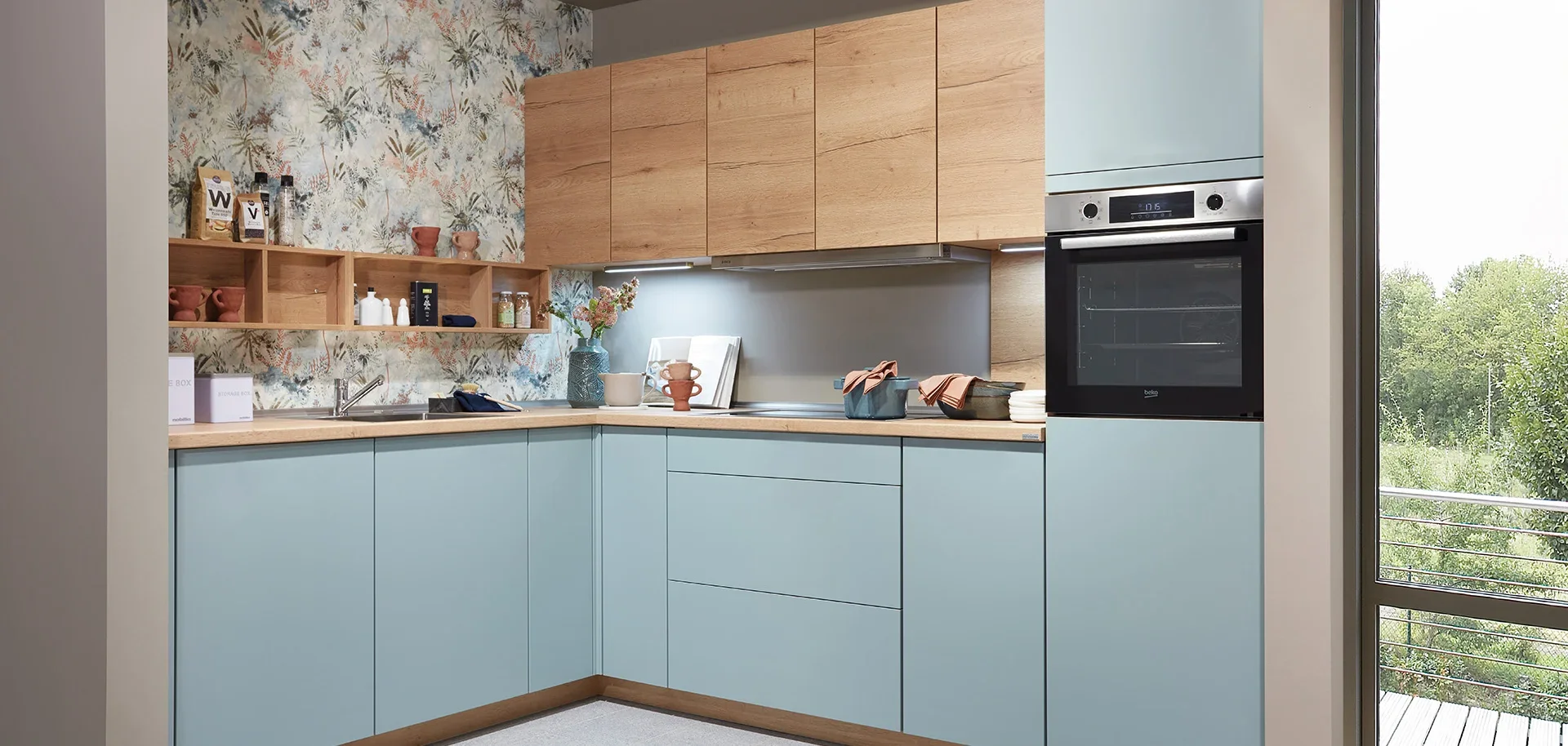 Modern kitchen with blue cabinets, wooden countertops, and a patterned backsplash with botanical designs. Open shelving displays jars, cups, and decorative items. A built-in oven and microwave are incorporated into the cabinetry, and a sliding glass 