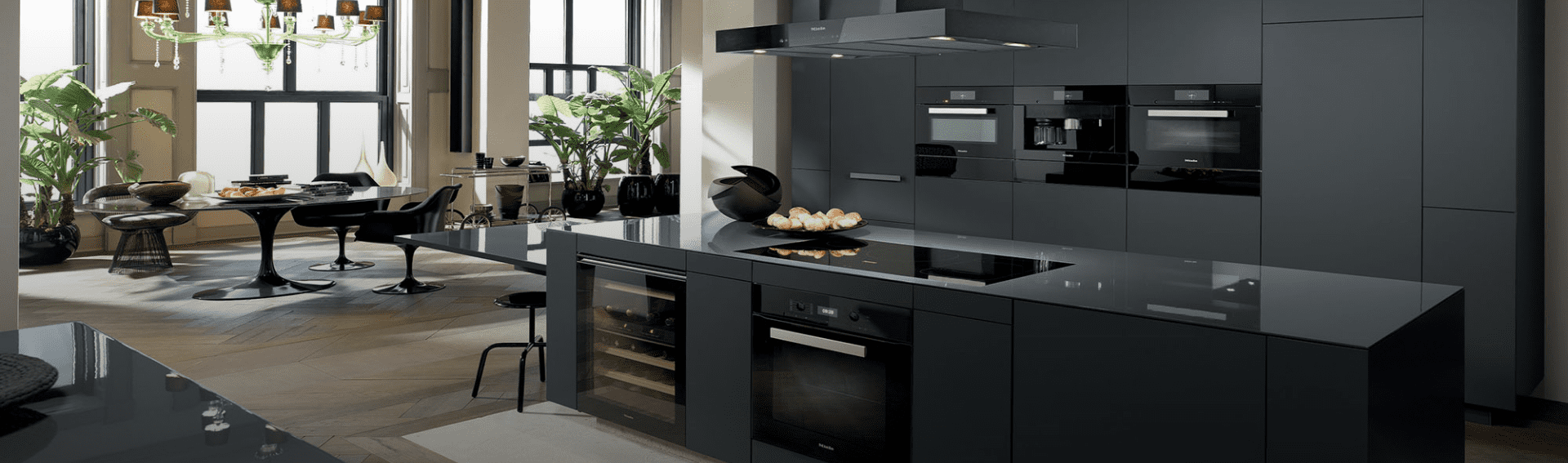 Modern black kitchen with built-in appliances, black countertop, and decorative black bowls, with a dining area featuring a black table and chairs, large windows, and indoor plants.