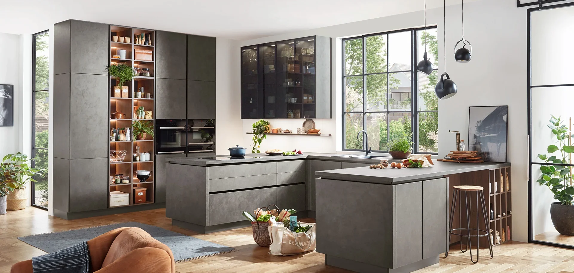 Modern kitchen with gray cabinets, large windows, black pendant lights, and wooden accents, decorated with plants and kitchenware.