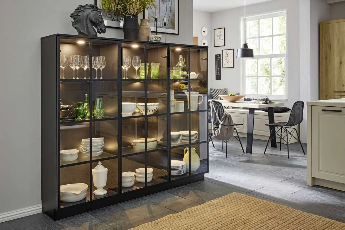 A black display cabinet with glass doors containing glassware, bowls, and decorative items in a modern dining area, with a window and chairs in the background.