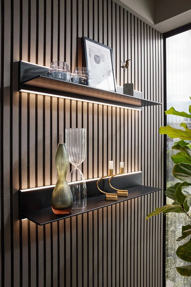 Decorative shelves with glassware, framed artwork, a small sculpture, and candles, against a wooden slat accent wall near a window with a green plant nearby.
