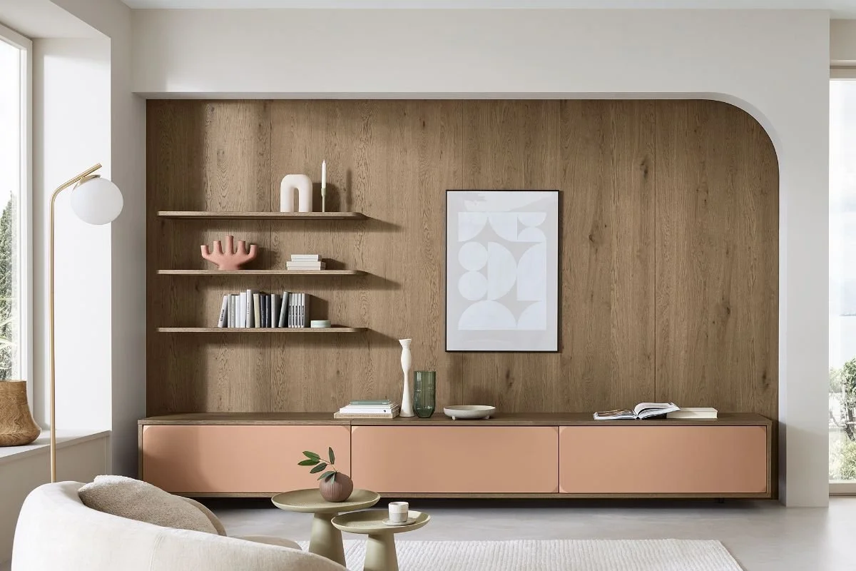Living room with a wooden wall, pink storage unit, minimalist decor, framed art, floating shelves with books and decorative objects, window with natural light, beige couch, and a white rug.