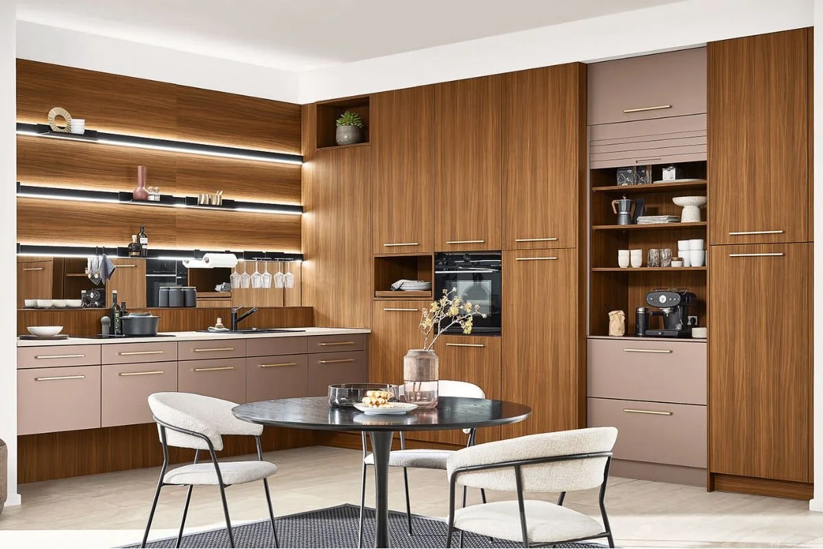 Modern kitchen with wooden cabinets, beige lower cabinets, and a black dining table with three beige chairs. Open shelving with decorative items, built-in oven, and coffee machine on the right wall.