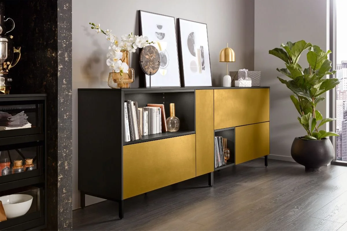Modern living room with a black and gold sideboard, abstract artwork, a gold table lamp, and a large potted plant near a window.