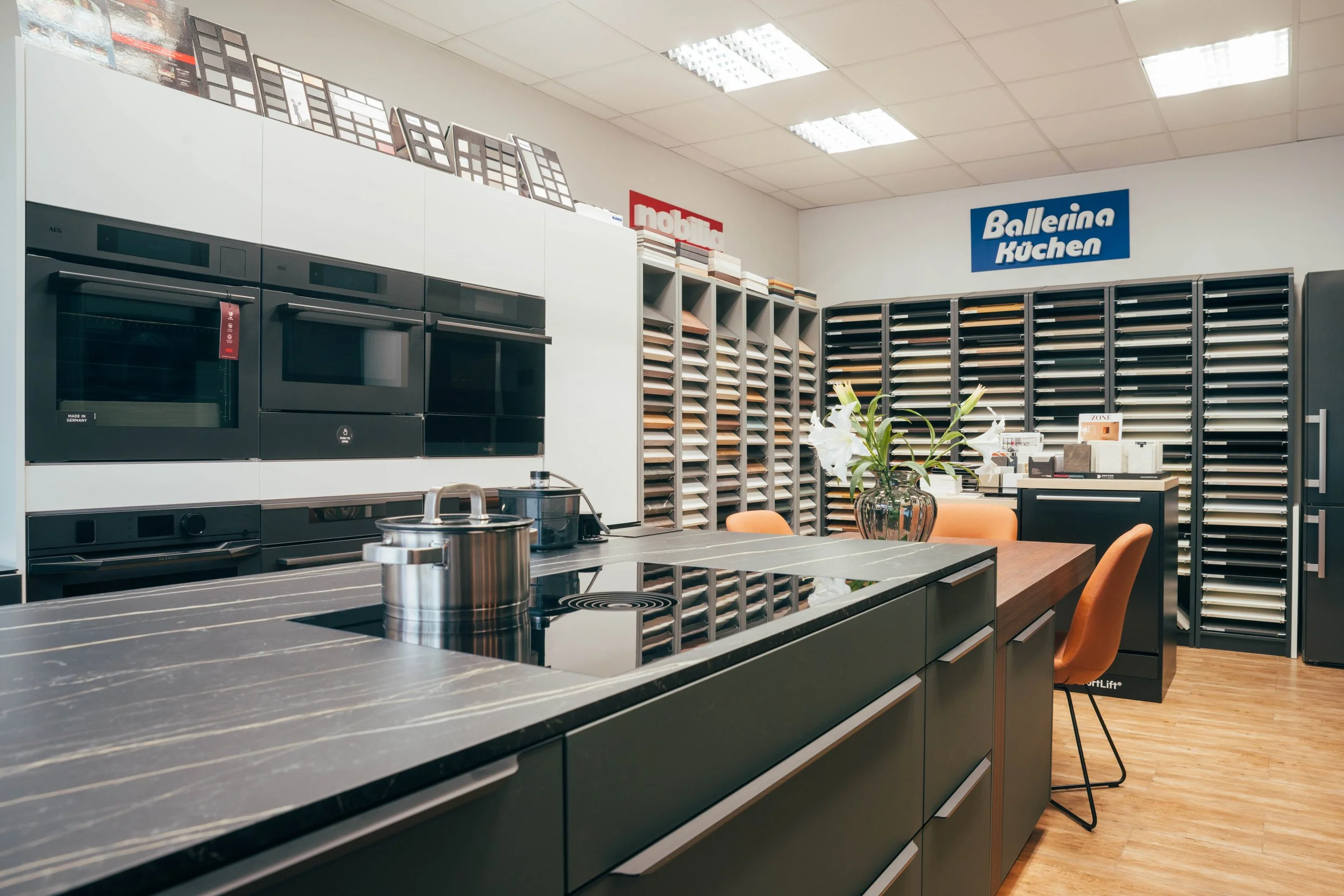 Showroom with kitchen appliances, sample cabinets, and decor. A black kitchen island with a pot and a skillet, orange chairs, and signage with brand names like Ballerina Küchen and Nobilia.