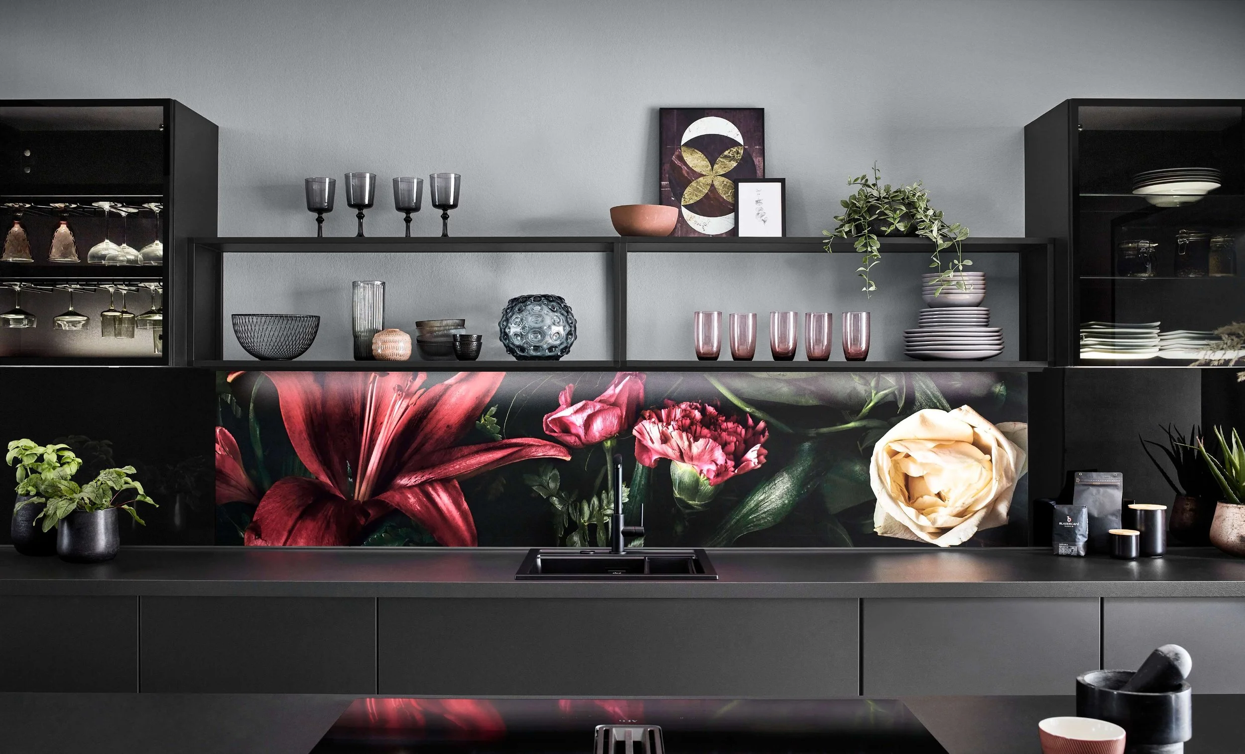Modern kitchen with black cabinets, a floral backsplash, and open shelving displaying glasses, bowls, and plants.