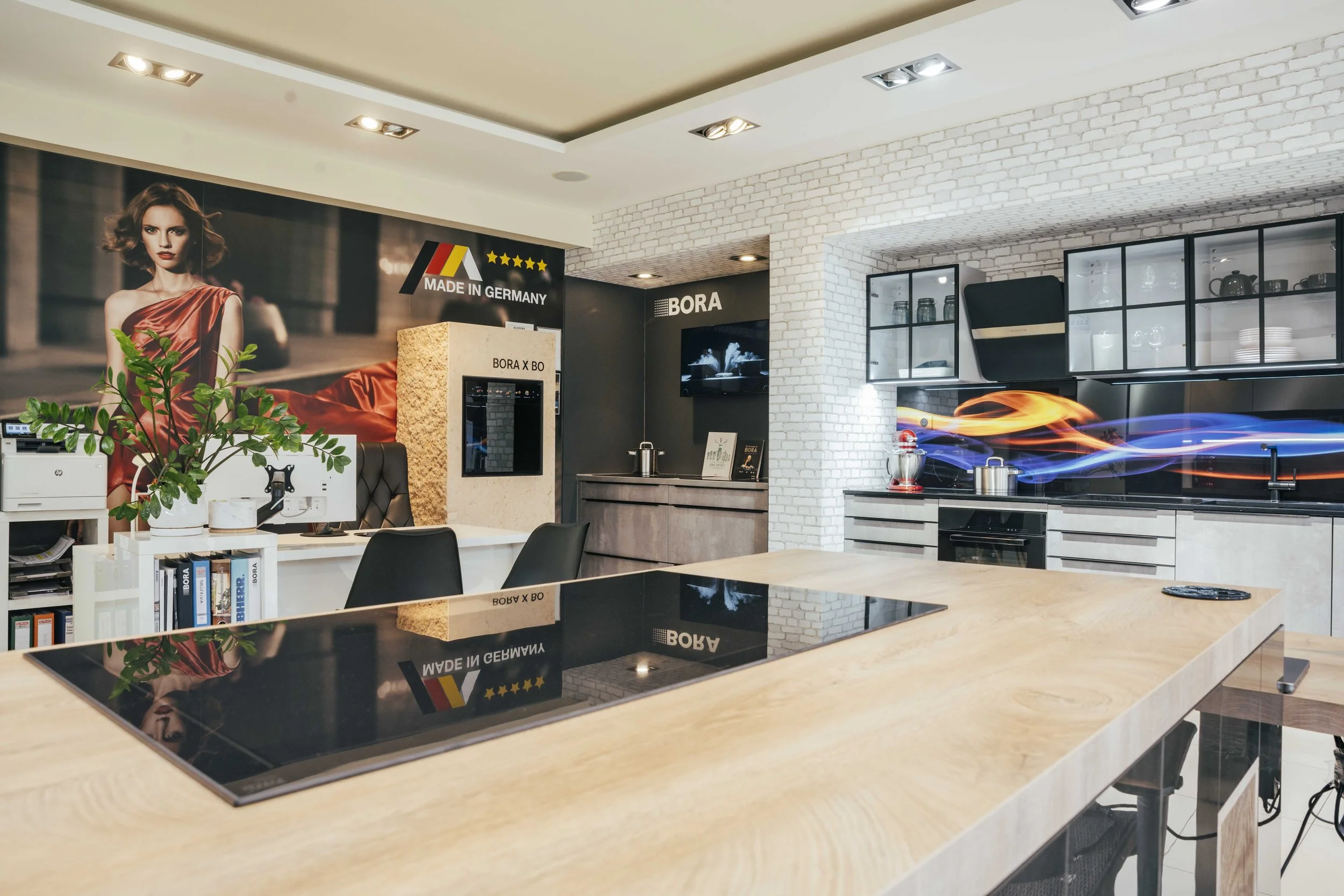 Modern kitchen with white brick walls, black cabinetry, a large black countertop with a built-in stove, glass-front cabinets, and a wall-mounted TV.