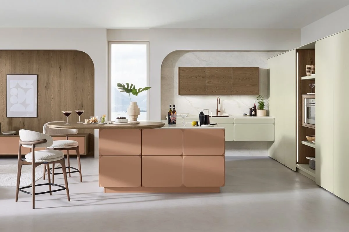 Modern kitchen with light green cabinets, wooden upper cabinets, a pink kitchen island, and a breakfast bar with two chairs. There is a window behind the island, some wine bottles, a vase with a large leaf, and various kitchen items.