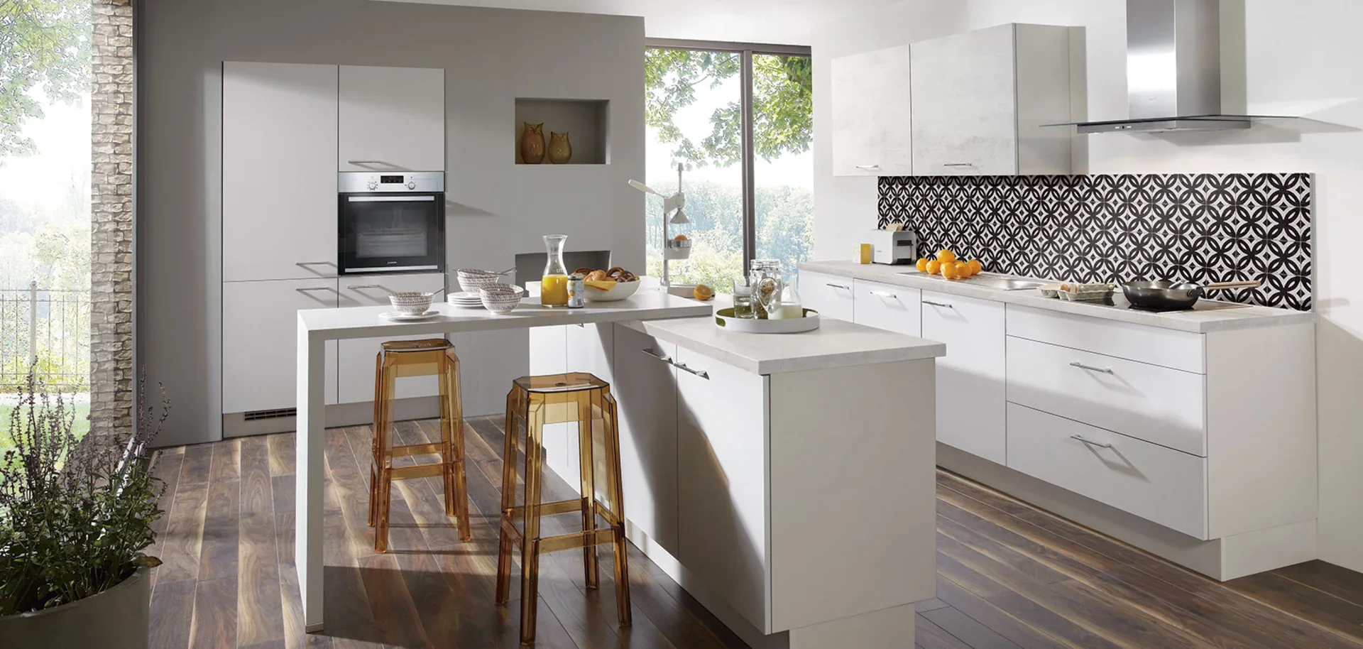 Modern kitchen with white cabinets, black and white patterned backsplash, wooden floor, large window with outdoor greenery, kitchen island with barstools, appliances, and breakfast setup