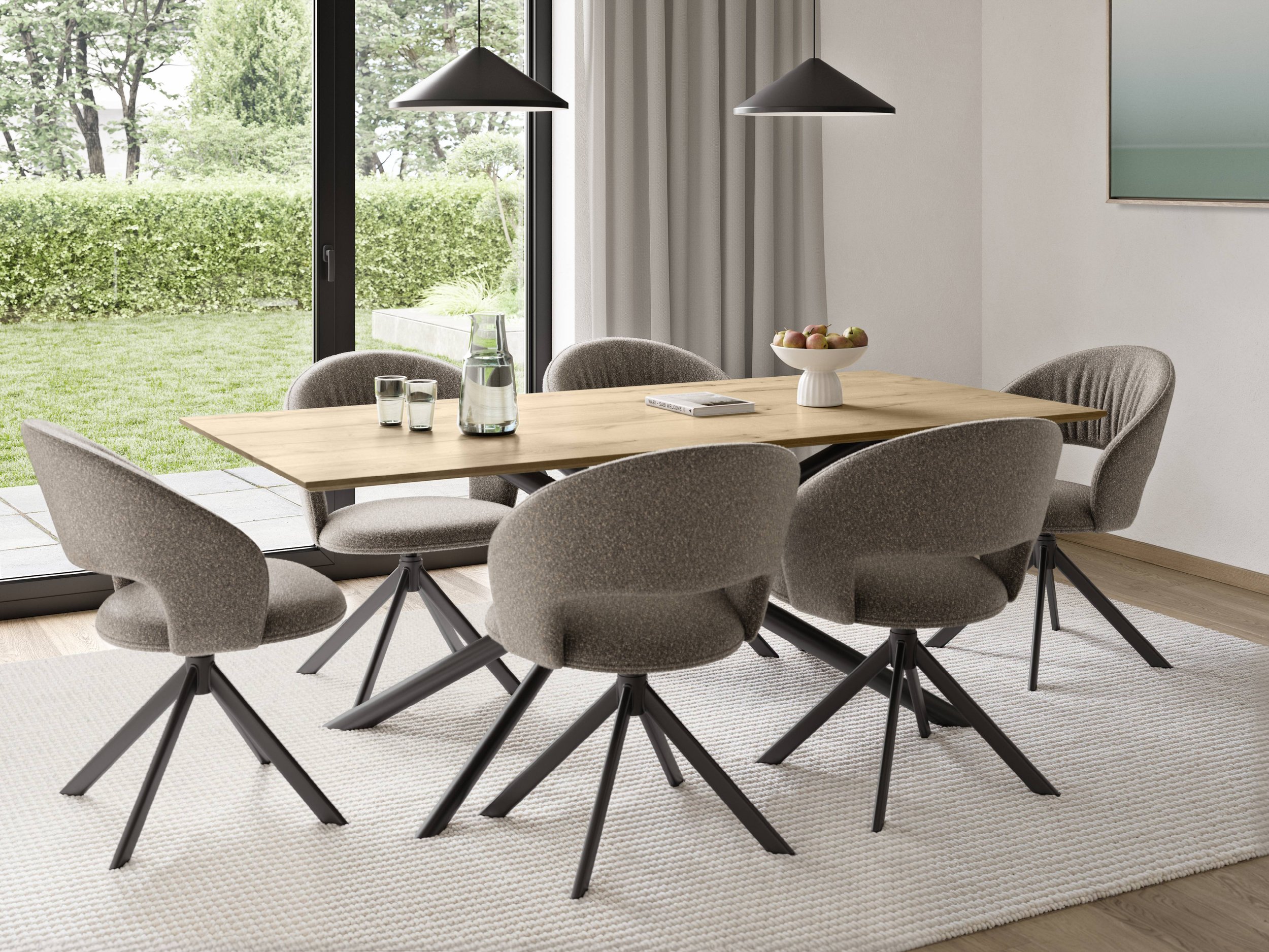 A dining room with a wooden table, six upholstered chairs, a glass pitcher and glasses, a white bowl of apples, and pendant lights, with large sliding glass doors leading to a green outdoor yard.