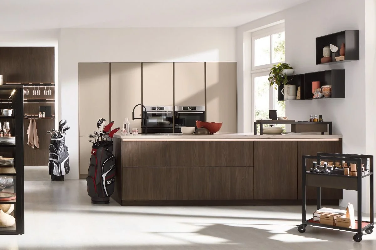 Modern kitchen with white walls, wooden cabinets, and large window letting in natural light. Two golf bags with clubs are placed next to the kitchen island. The kitchen features built-in ovens, open shelving with decorative items, and a black cart wi