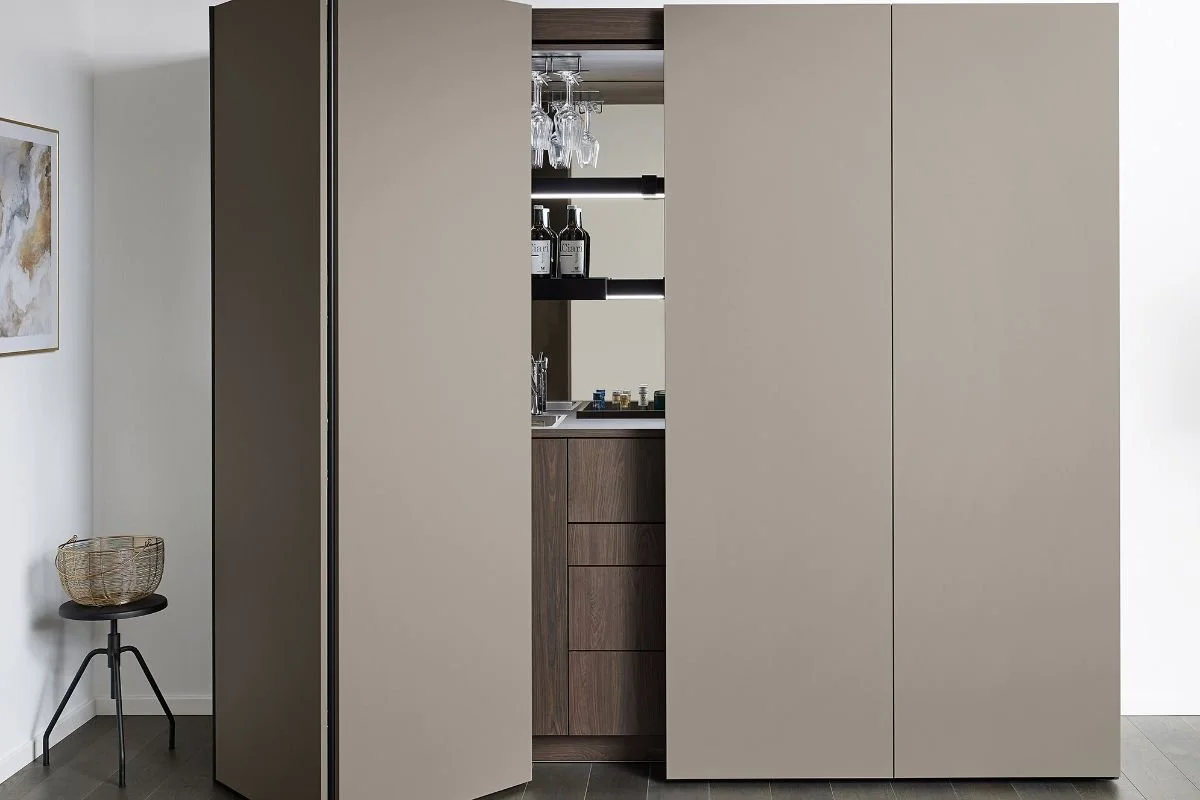 A modern, beige-colored folding door revealing a small wet bar area with wine glasses hanging upside down and bottles of alcohol on shelves, adjacent to a closet or storage space.