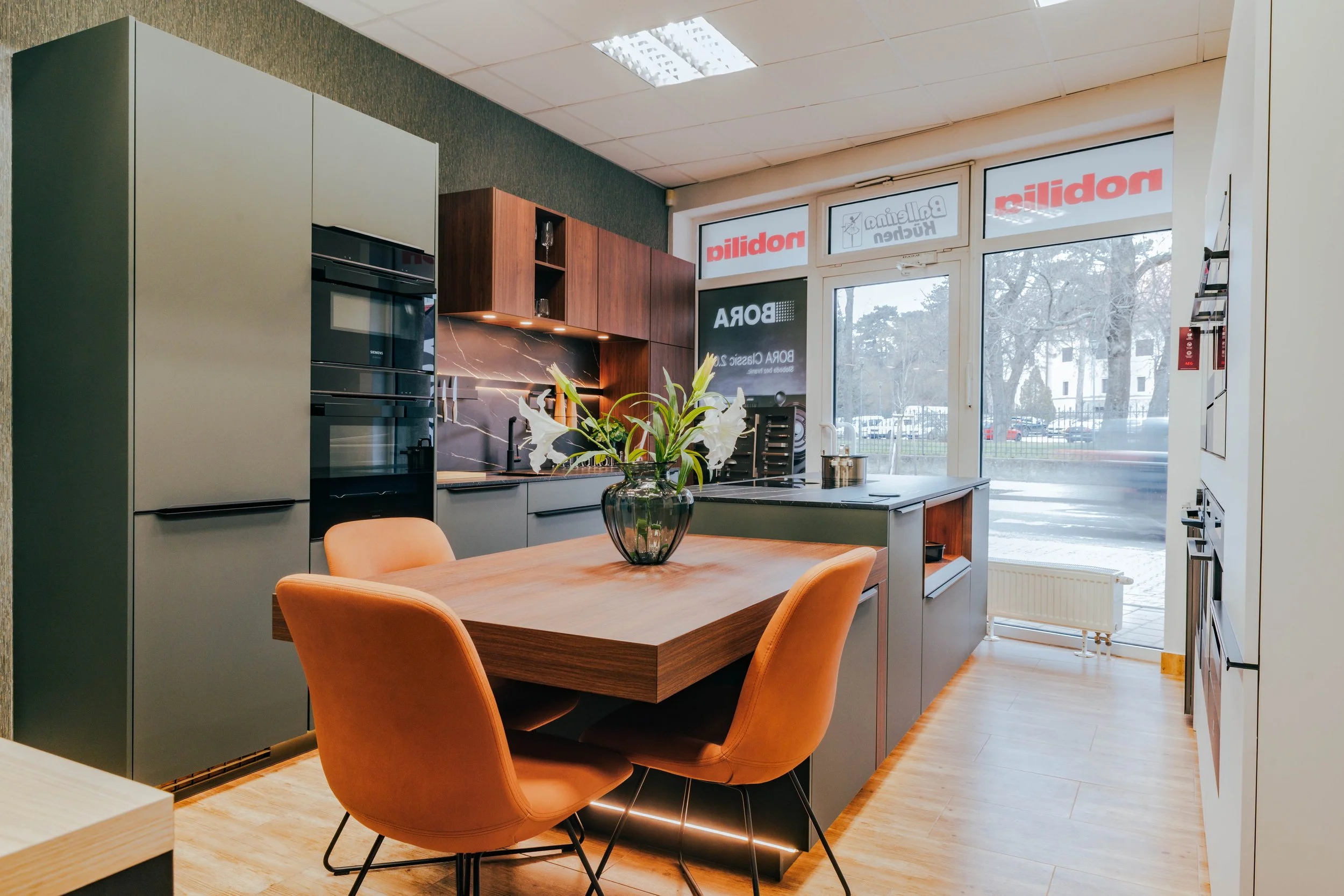 Modern kitchen with gray cabinets, wooden accents, a black marble backsplash, and a window with red and white signs. There is a wooden table with orange chairs and a vase of white lilies.