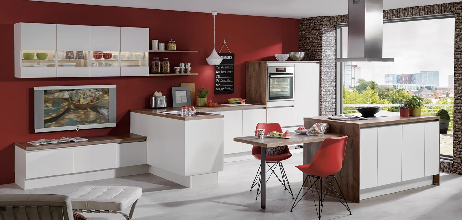 Modern kitchen with white cabinets, a red accent wall, and a large window overlooking a cityscape with trees. Countertop with bowls, a plant, and food items. Small table with red chairs. TV on the wall showing food, and various kitchen appliances and