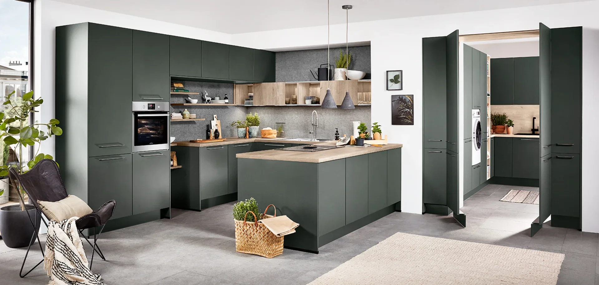 Modern kitchen with green cabinets, open shelving, built-in oven, and a kitchen island with plants and kitchen utensils. There is a laundry area with washer and dryer tucked in a folding screen.