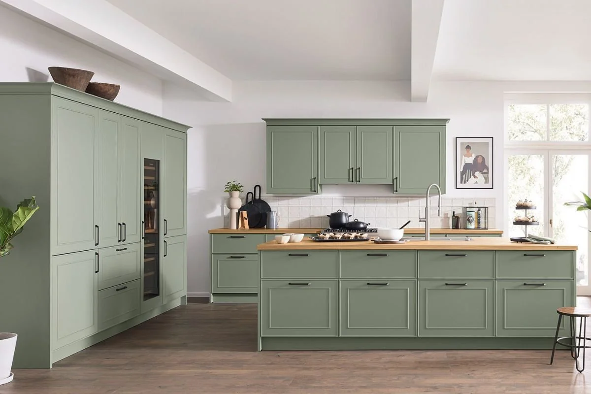 Modern kitchen with sage green cabinets, wooden countertops, and a white backsplash. There is a black stove with a pot, a sink, and some decorative items like a black handbag and framed artwork.