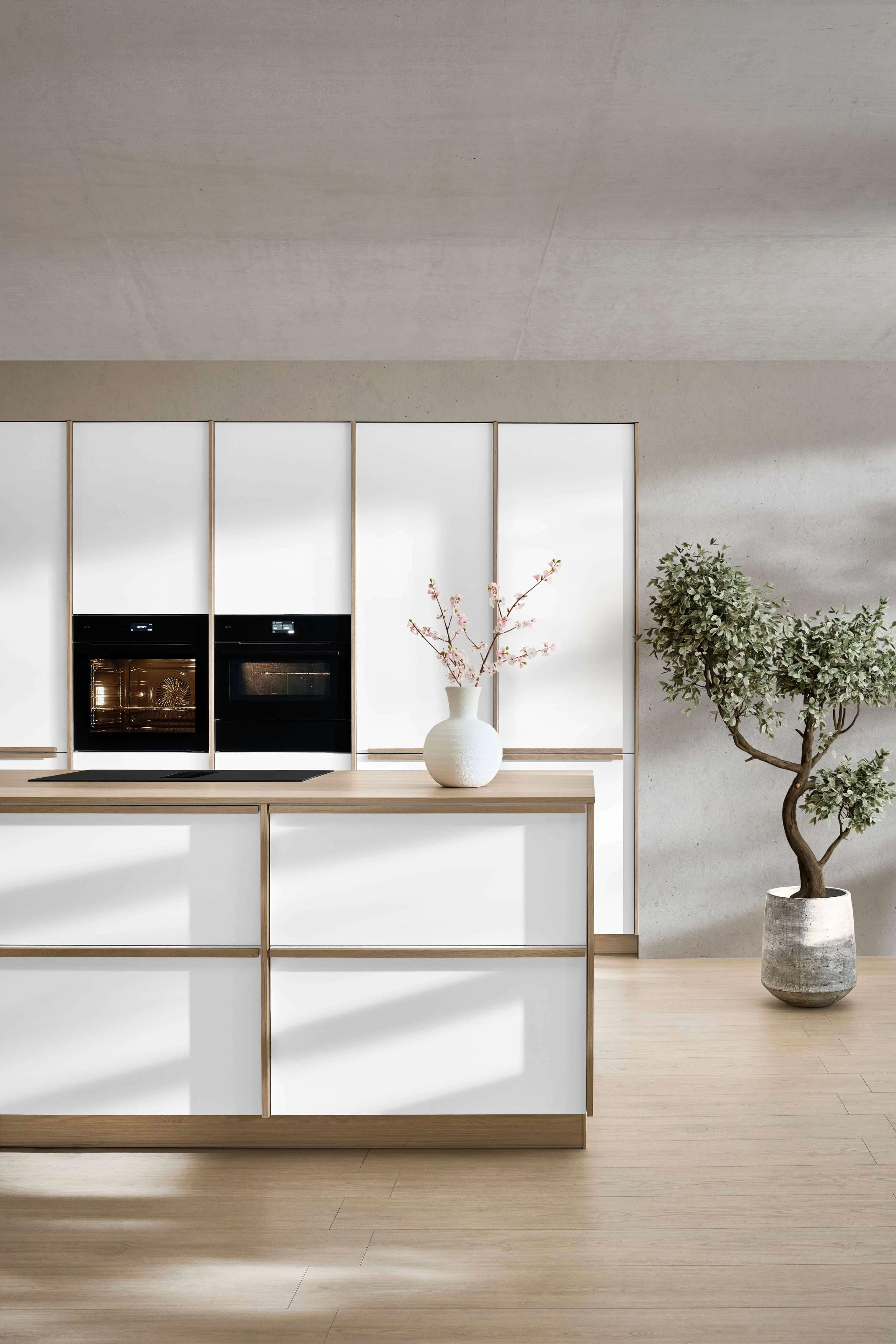 Modern kitchen with built-in black oven, white cabinetry, a white vase with pink cherry blossom branches, a large potted green plant, light wood floors, and abundant natural light.