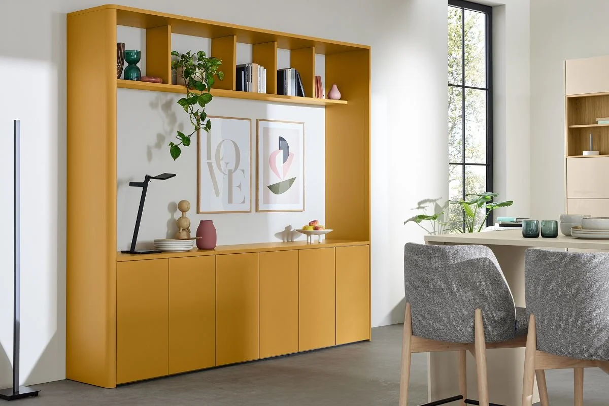 Modern dining and living area with a yellow built-in storage unit, framed art, decorative objects, and a gray upholstered chair next to a round dining table with green glassware and white dishes, large window with greenery outside.