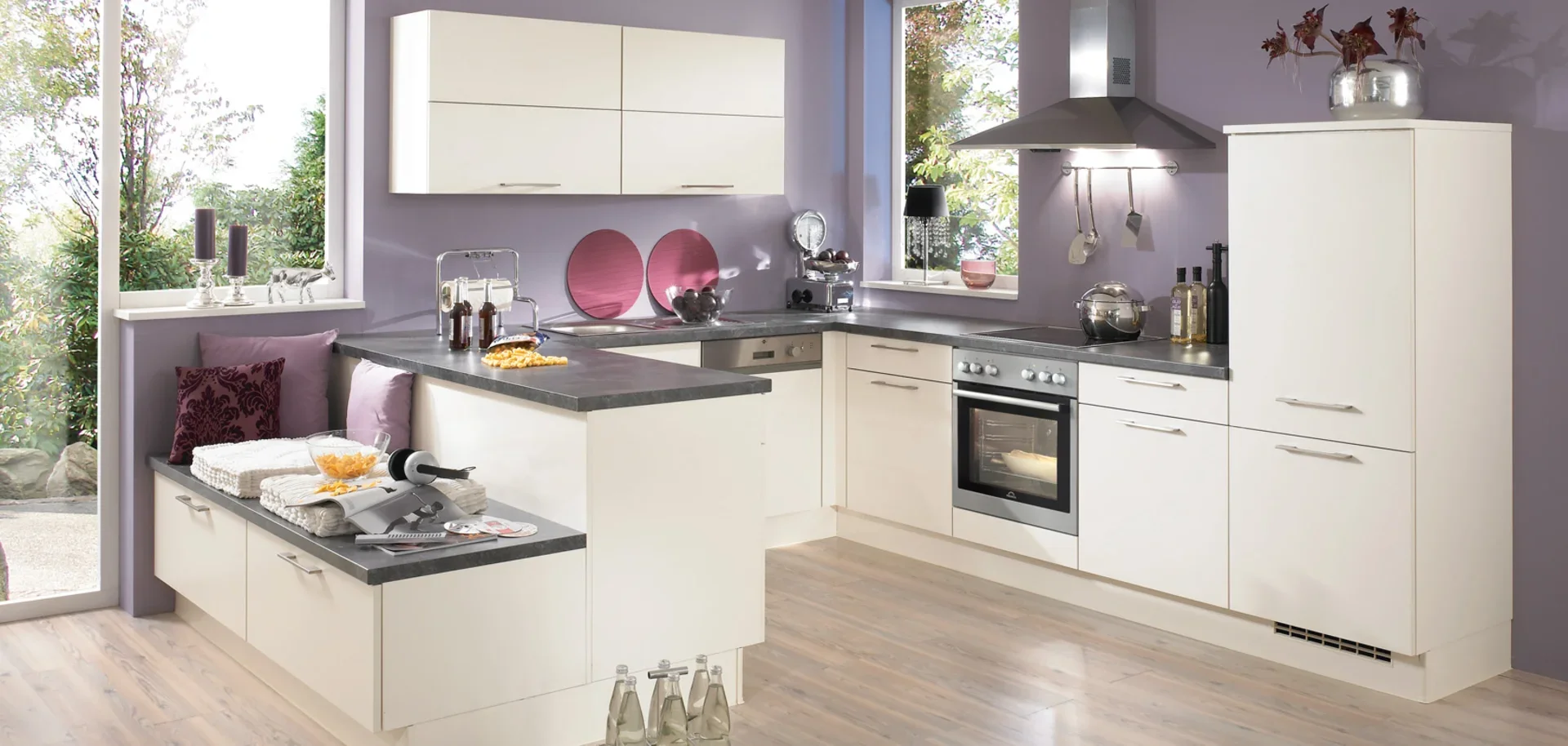 Modern kitchen with white cabinets, grey countertops, purple walls, and large windows showing outdoor greenery.