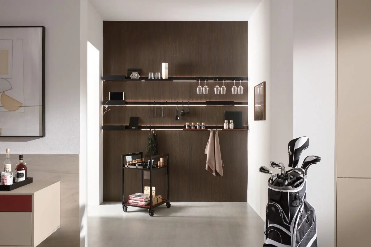 Inside a modern room with a dark wooden wall, multiple shelves hold glassware, tablets, jars, and decorative items. To the right, a golf bag with clubs stands next to a beige wall, while on the left, a corner of a countertop with bottles. There's art