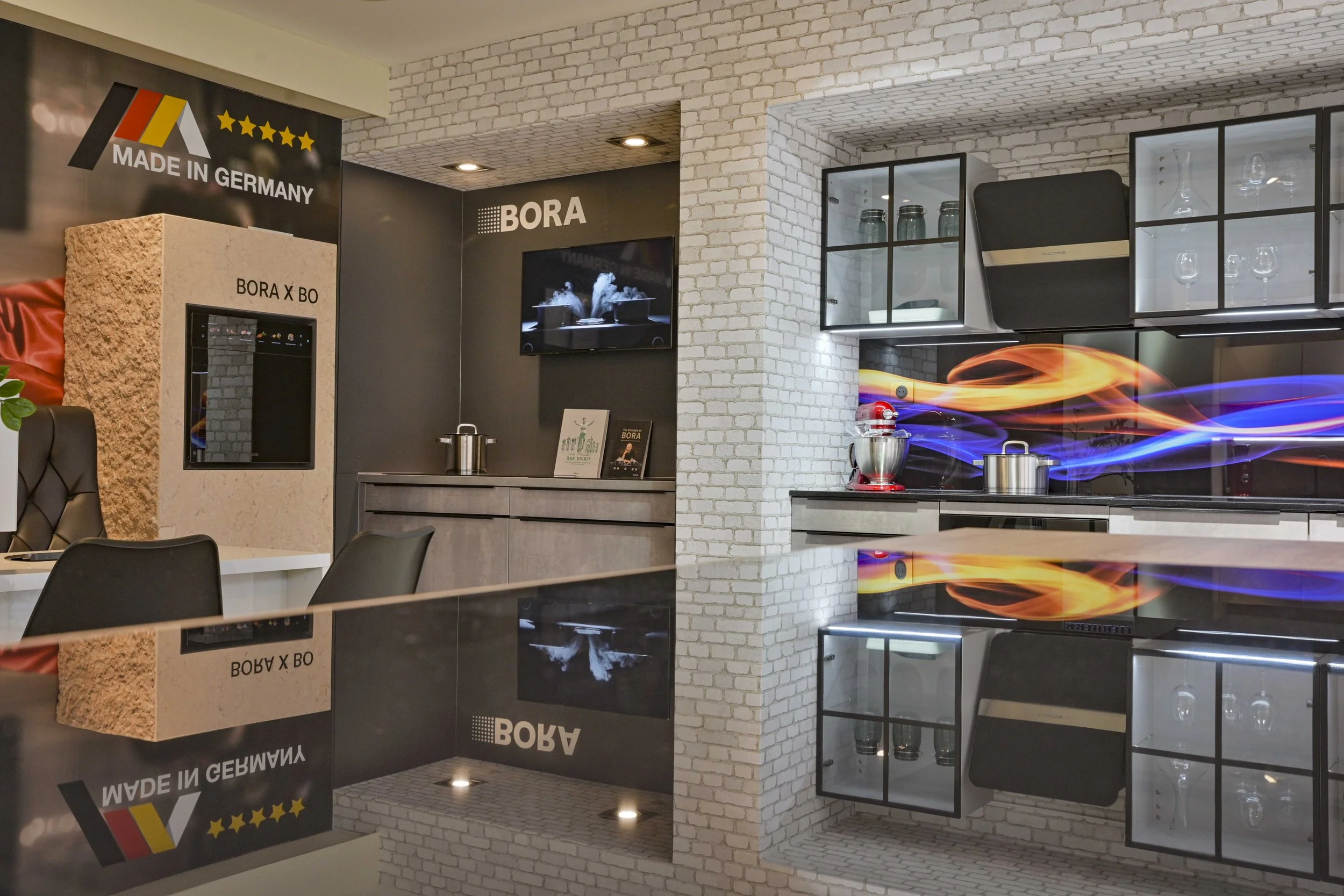 Modern kitchen display area with black cabinets, a white brick wall, glass-fronted cabinets with glassware, and a TV screen showing steam rising from bowls. There are colorful flames or smoke effects in the foreground, and a sign that reads 'MADE IN 