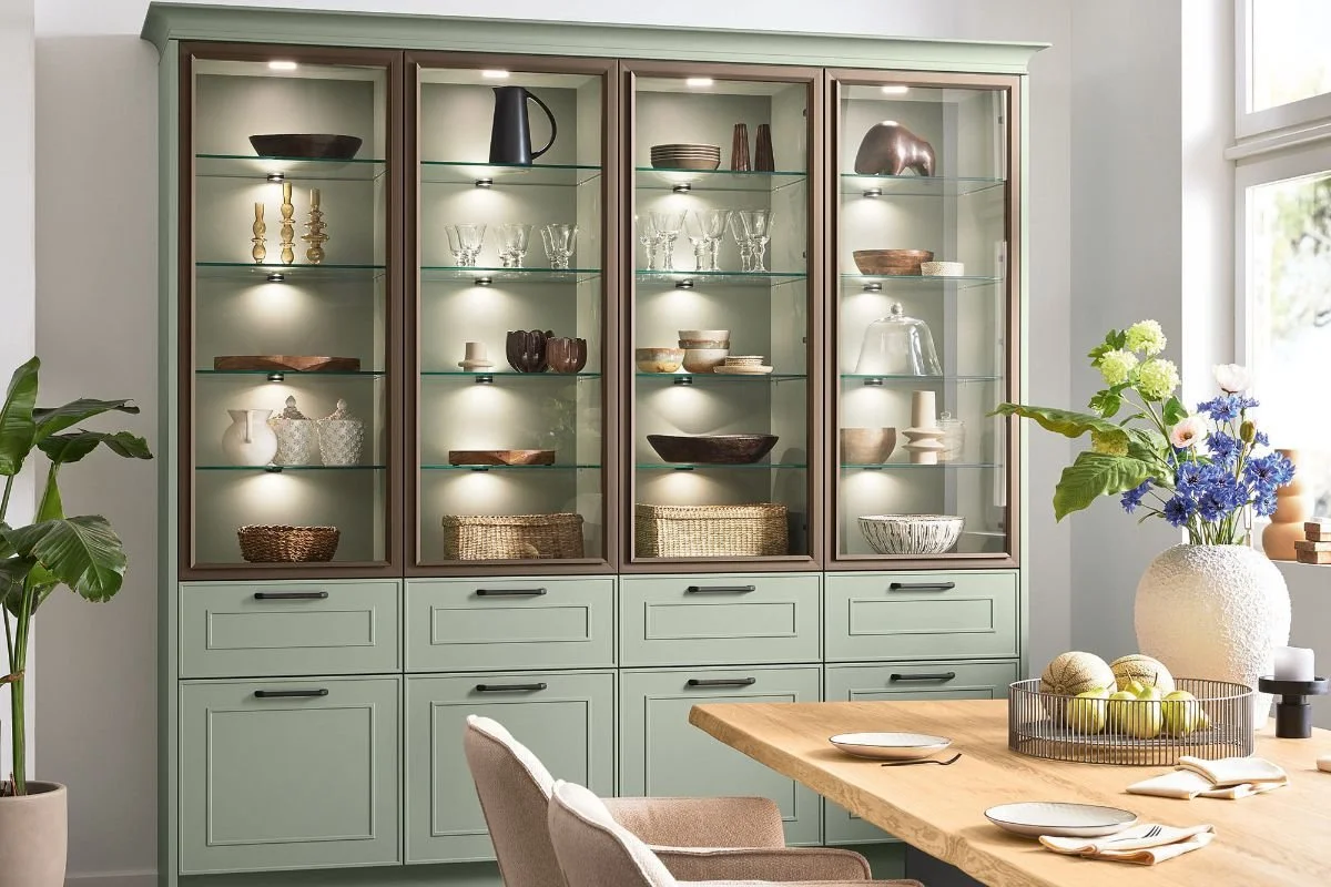 A light green and brown cabinet with glass doors displaying various glassware, bowls, and decorative objects in a bright dining room with a wooden table, chairs, a large vase of flowers, and a window.