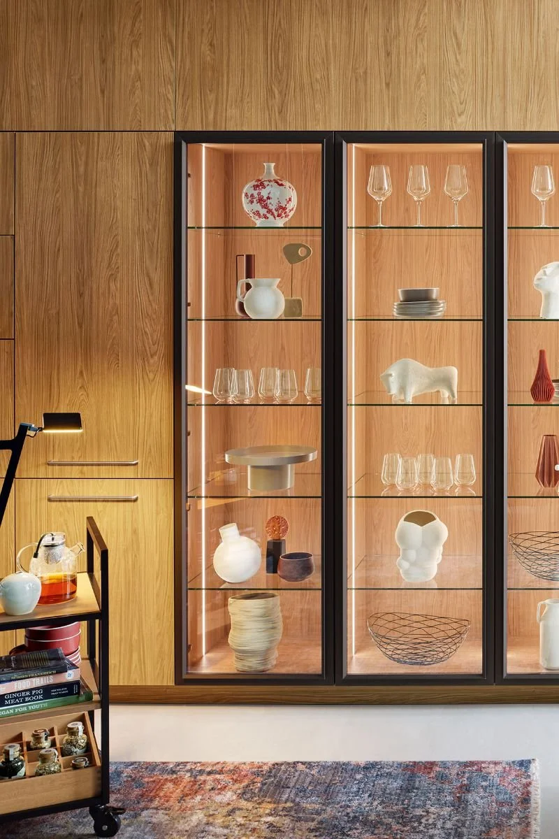 A wooden display cabinet with glass doors showcasing various decorative ceramics and glassware in a modern interior setting.