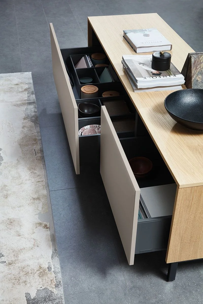 A wooden dining table with open cabinet drawers underneath, containing dishes and bowls, with books, a black container, and decorative items on top, situated on a tiled floor next to a light-colored rug.