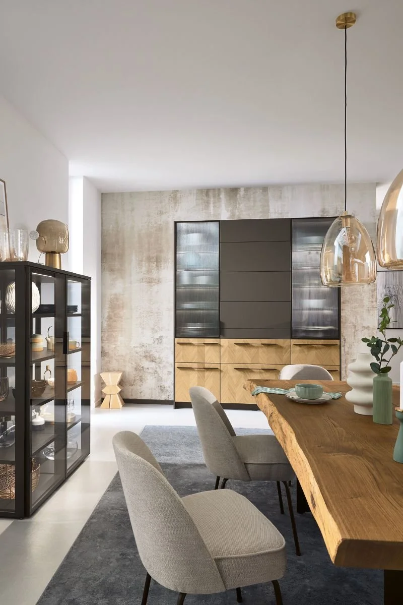 Modern dining room with a natural wood table, beige upholstered chairs, black display cabinet, and a gray textured wall with a black and wood storage unit. Decor includes vases, artworks, and hanging light fixtures.
