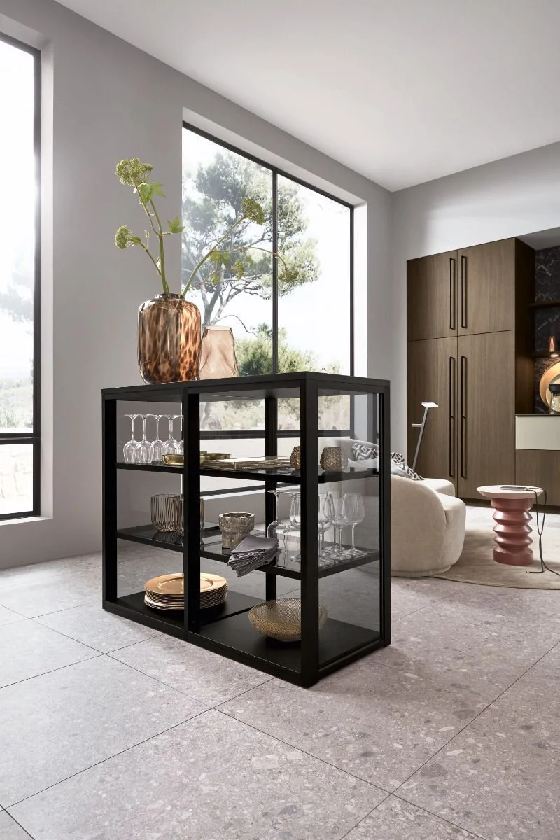 Modern living room with large windows, a black display shelf with glassware and decor, a beige sofa, a pink side table, and wooden cabinets in the background.
