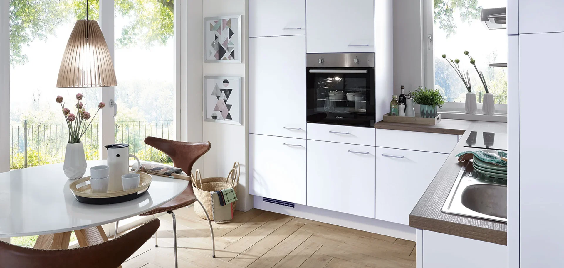 Modern kitchen with white cabinets, a built-in oven, a wooden countertop with a sink, and a small round dining table with a vase of flowers and cups, near large windows with a view of trees.