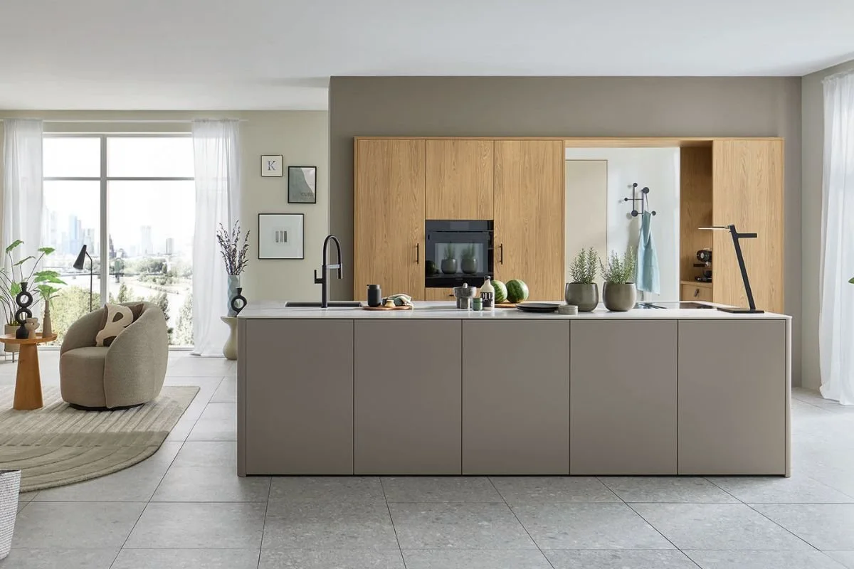 Modern, open-concept kitchen with beige cabinetry, a white kitchen island, gray flooring, and large windows with white curtains, overlooking a cityscape and greenery.