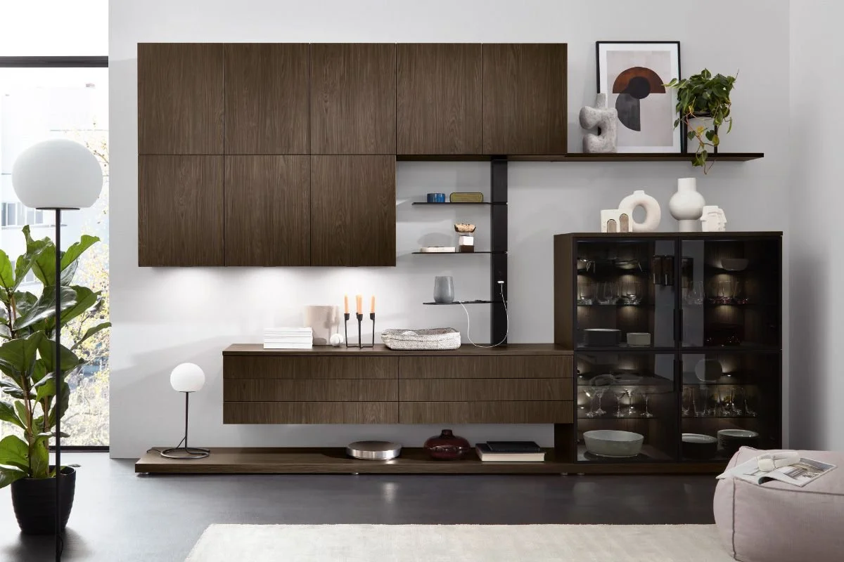 Modern living room with wooden cabinet wall unit, decorative art, and a black display cabinet with glass doors filled with glassware. A tall white floor lamp, a potted plant, and a small white table lamp are also present.