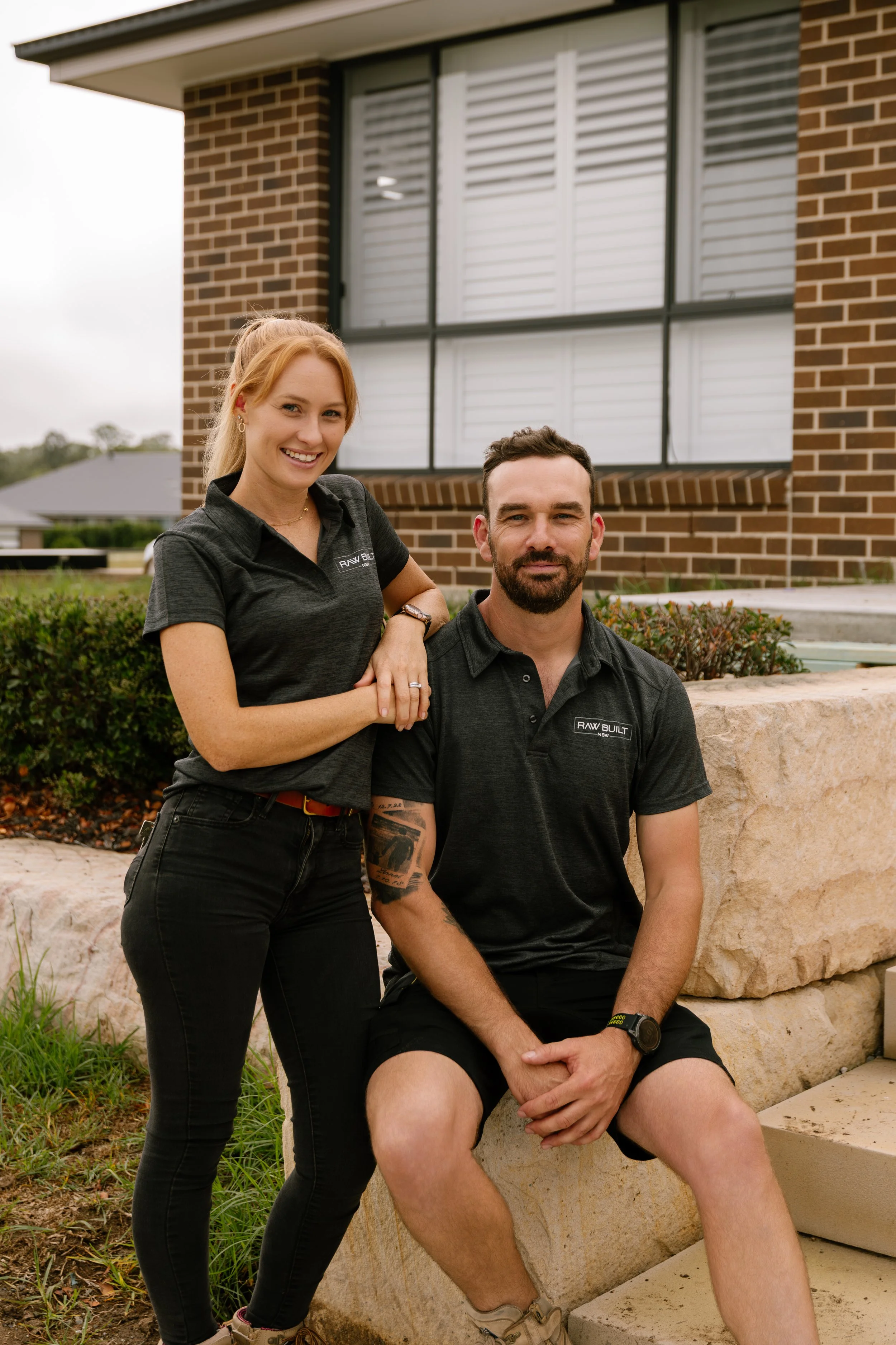 A man and woman posing outdoors in front of a brick house. The woman is standing and smiling, with one arm resting on the man's shoulder. The man is sitting on a stone ledge, looking at the camera with a neutral expression. Both are wearing black shirts with a logo that says "RAW BUILT." The woman has long, blonde hair, and the man has a beard and tattoos on his arm.