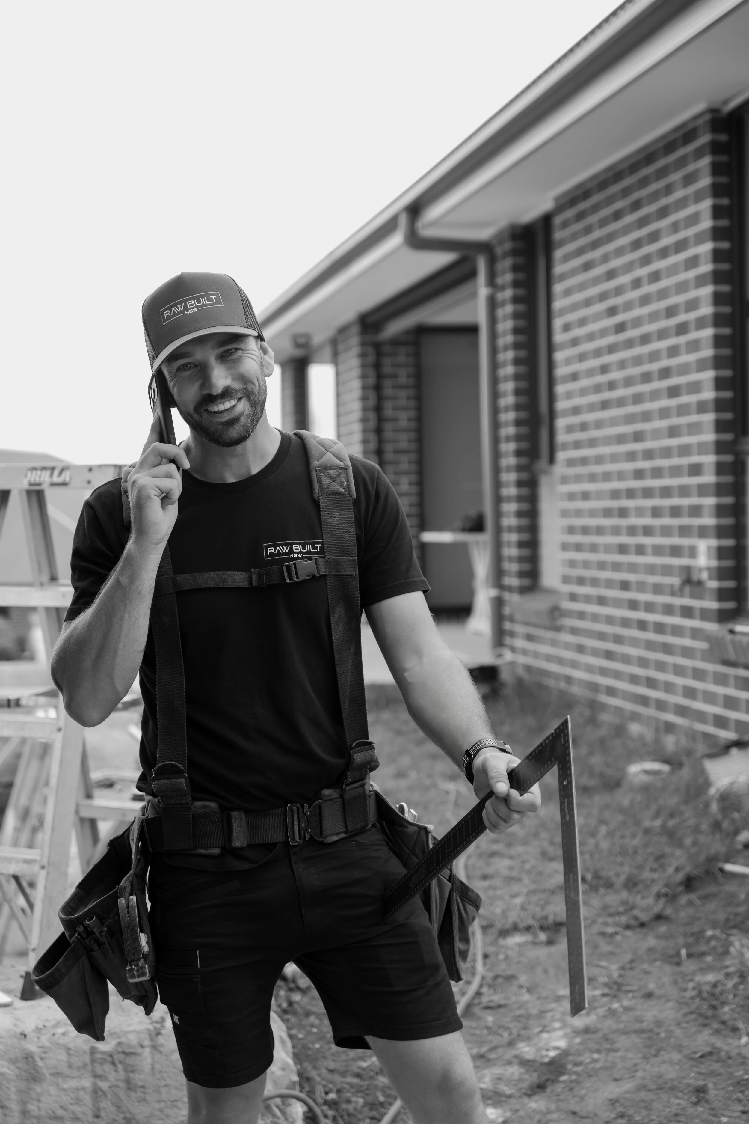 A smiling man with a beard, wearing a cap and a T-shirt that says 'RAW BUILT', is talking on a cellphone. He is holding a square-shaped tool or camera frame in one hand, and has a tool belt around his waist. The background features a brick building and some construction equipment.