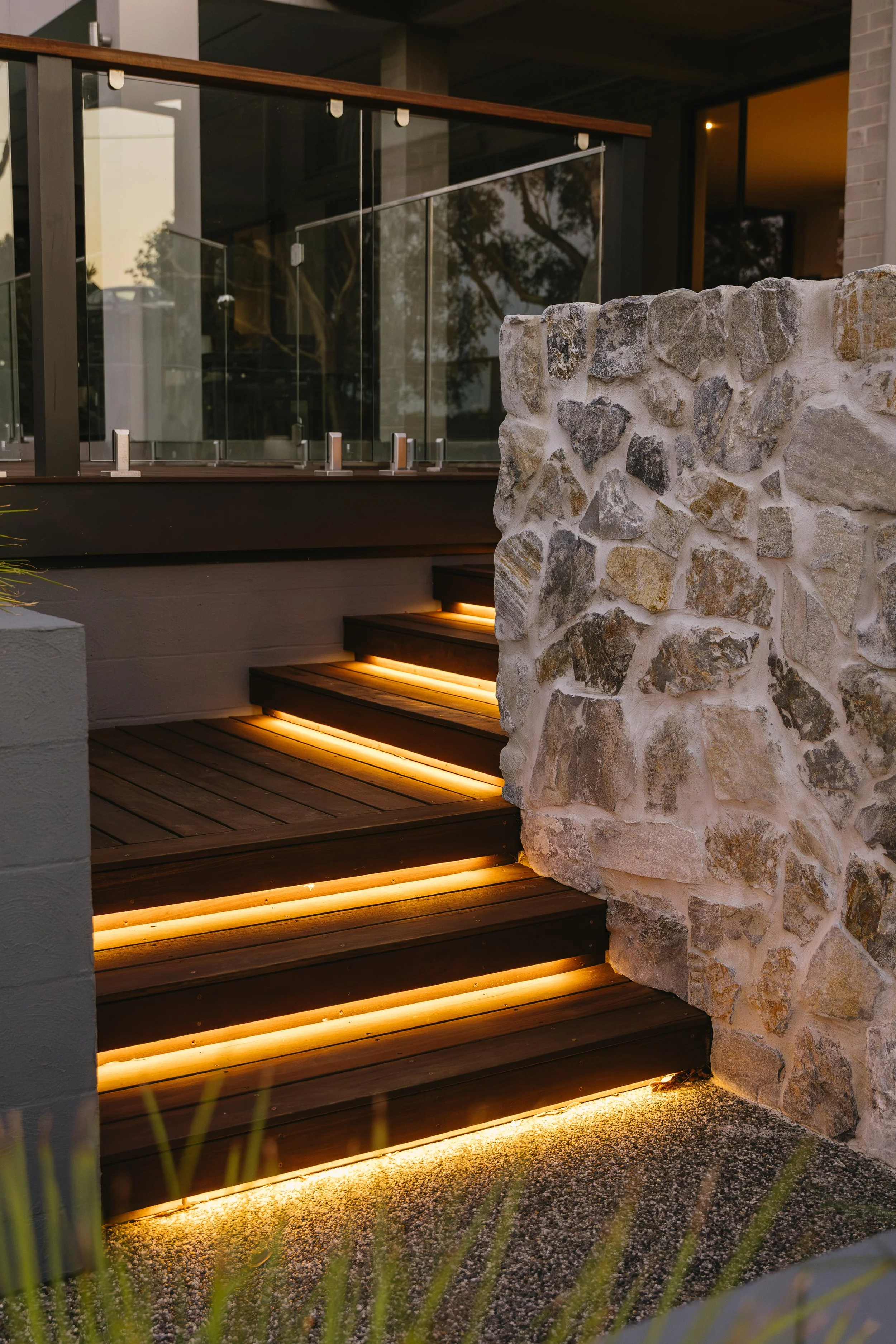 Exterior view of a modern building with a stone wall, glass railing, wooden stairs with integrated lighting, and reflected trees in the glass