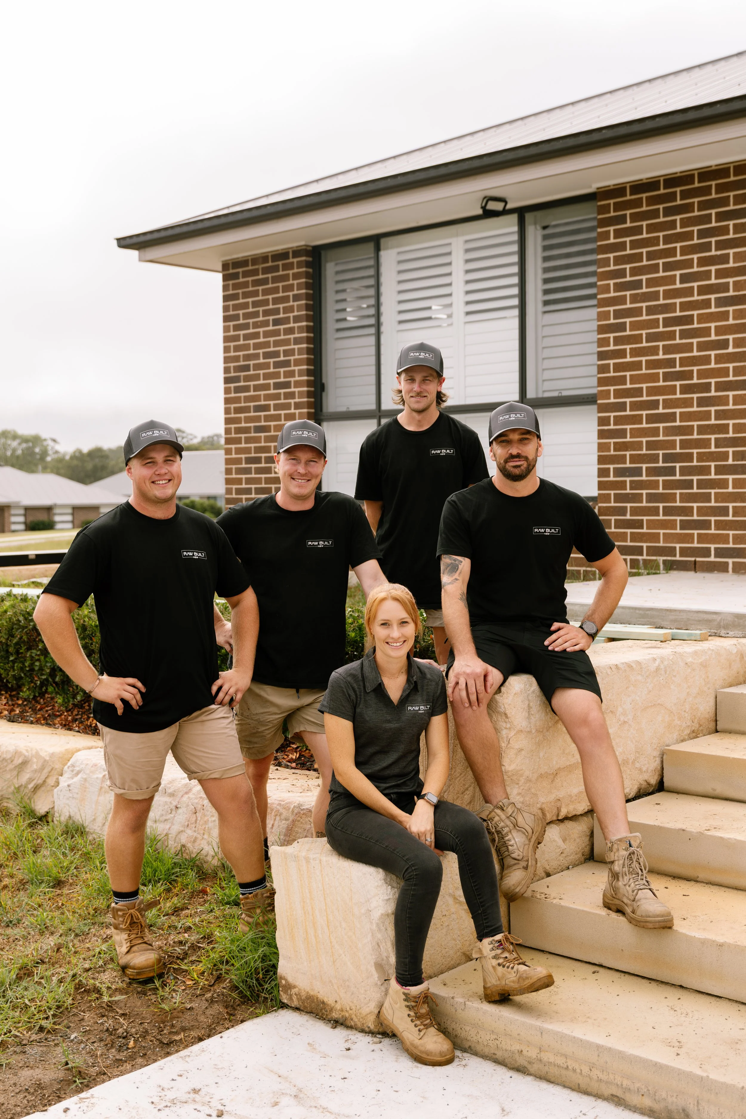 Group of five construction workers, four men and one woman, posing outdoors in front of a building with brick and large window, all wearing black shirts, beige or black pants, work boots, and black caps, smiling at the camera.