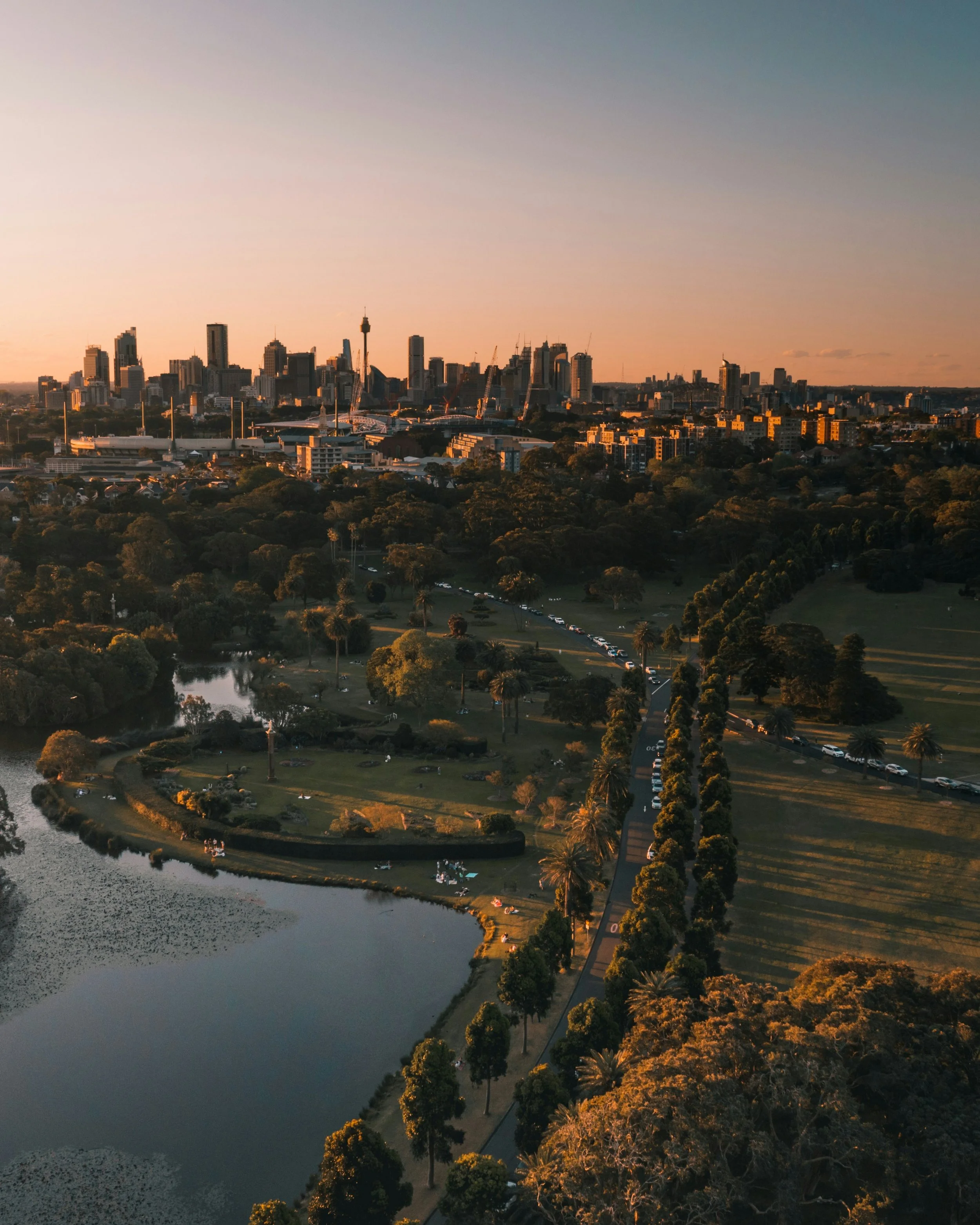 A city skyline at sunset viewed over Centennial park with trees, a pond, and a walking trail in the foreground.