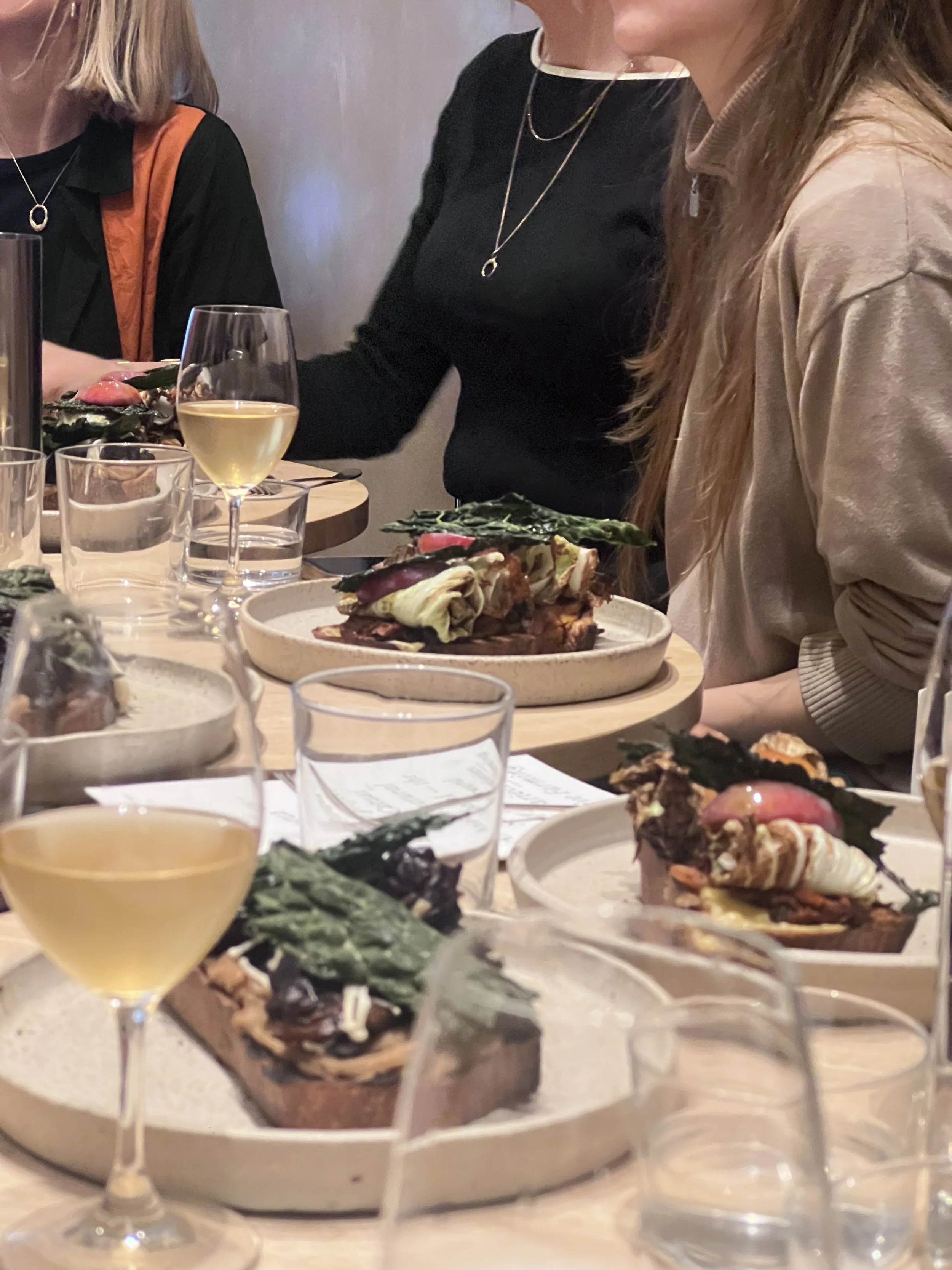 People sitting around a table with food and drinks, including plates of savory dishes topped with greens and seafood, and glasses of white wine.