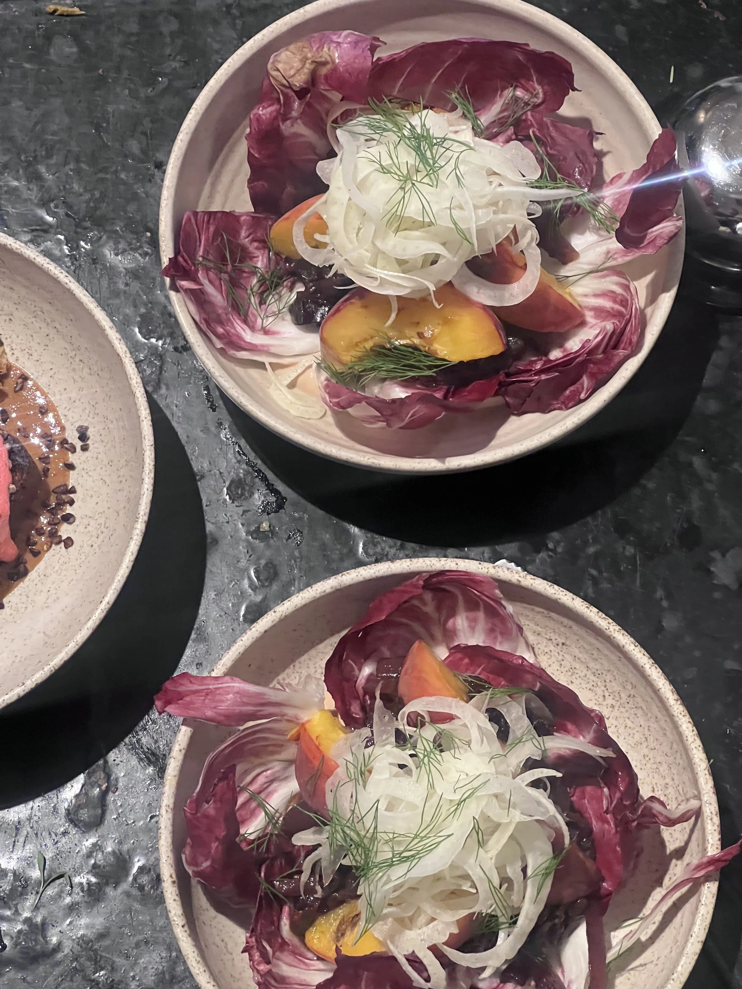 Two bowls of salad with radicchio, grilled peaches, fennel, and dill, on a dark table.