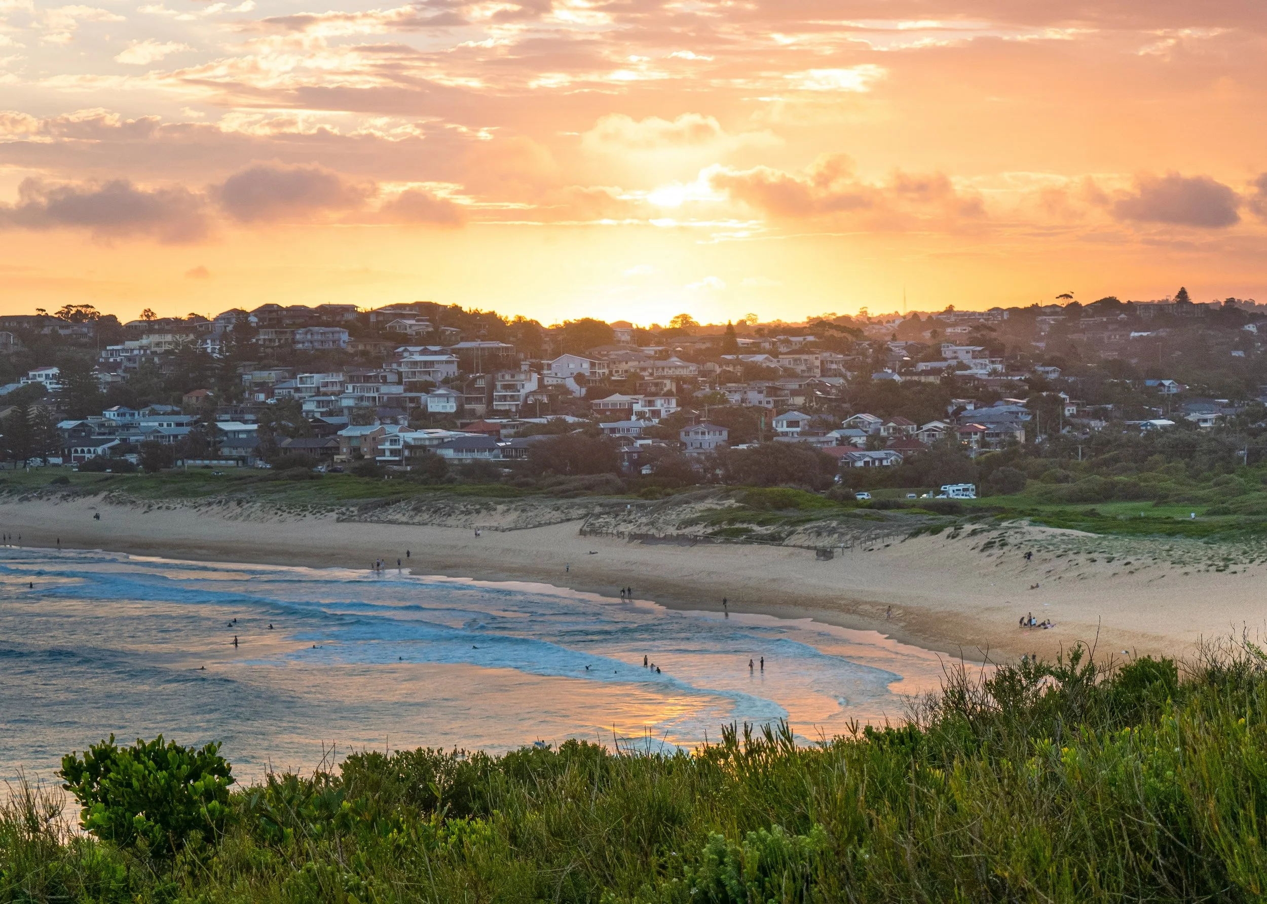 Sunset over Narrabeen beach with waves and people walking along the shoreline, with houses on a hillside in the background.