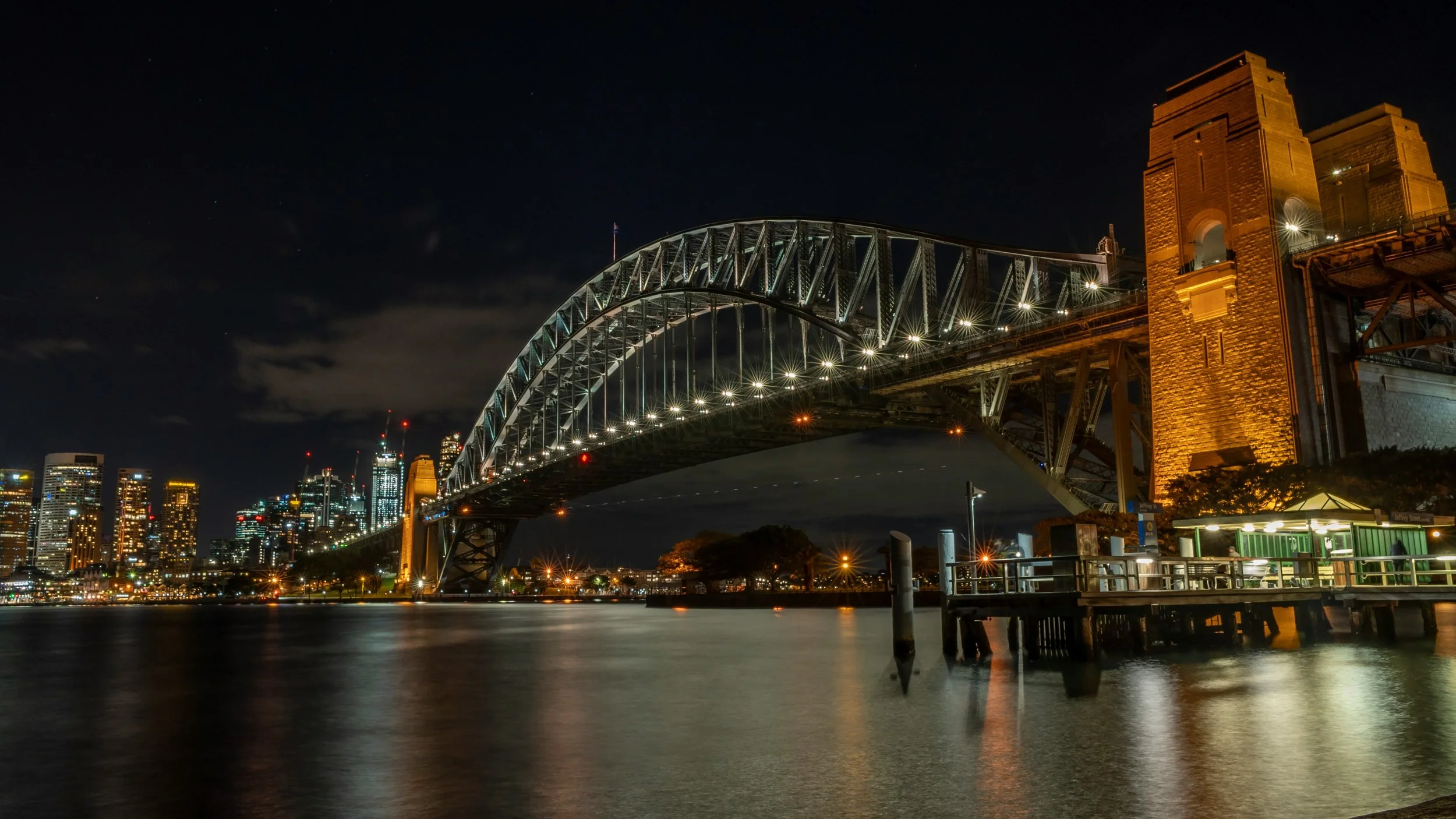 Night view of the Sydney Harbour Bridge with city skyline in the background, illuminated and reflected on the water.
