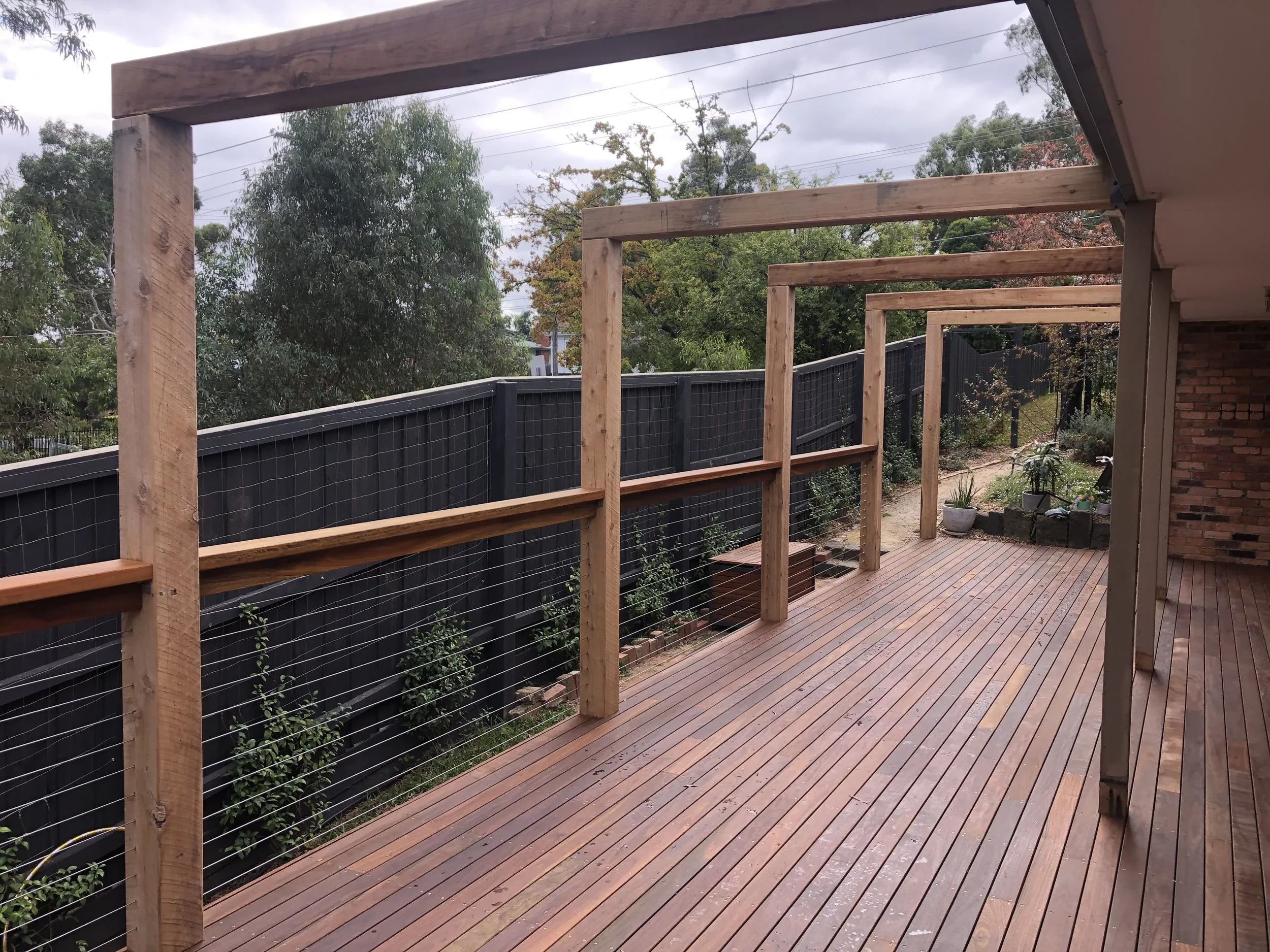 A wooden deck in the process of construction, with support beams and a top rail, overlooking a backyard with plants and a black fence, under cloudy skies.