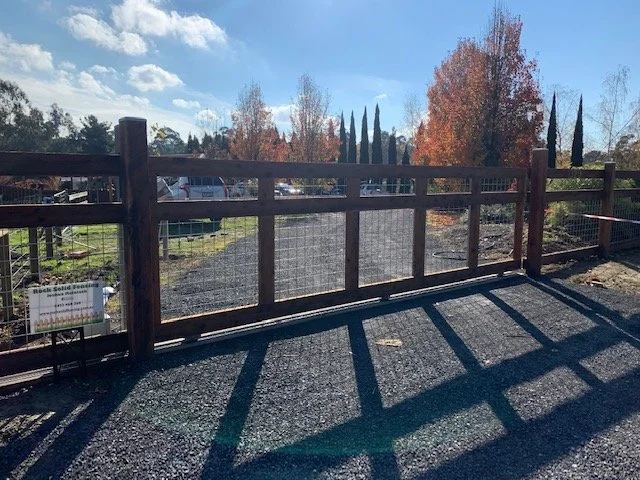 Wooden fence on a gravel lot with long shadows cast by the fence, trees with autumn foliage, and a partly cloudy sky.