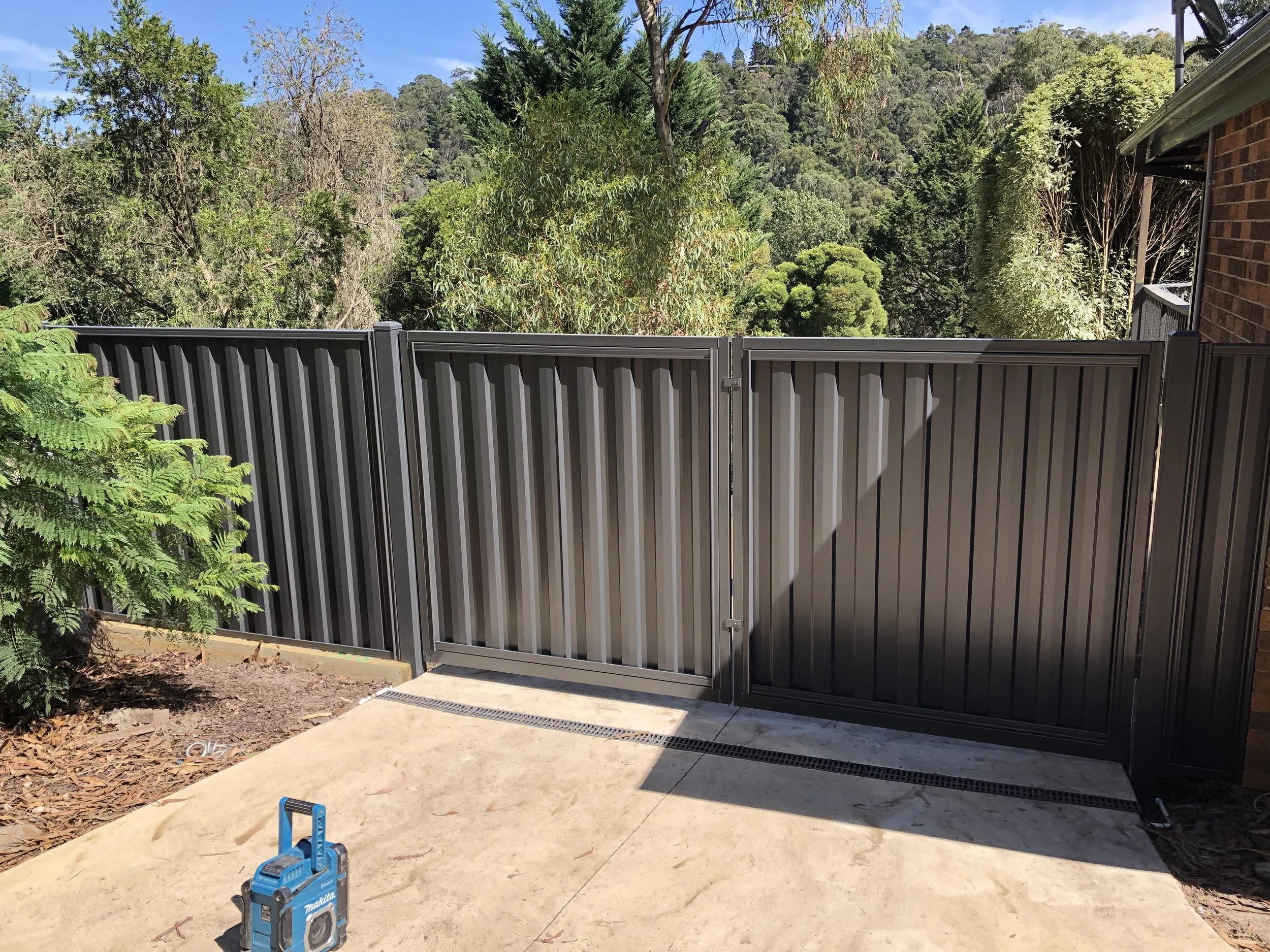 A metal gate and fence with a rural wooded landscape in the background, a small black device with a handle on the ground, and a brick building partially visible on the right.