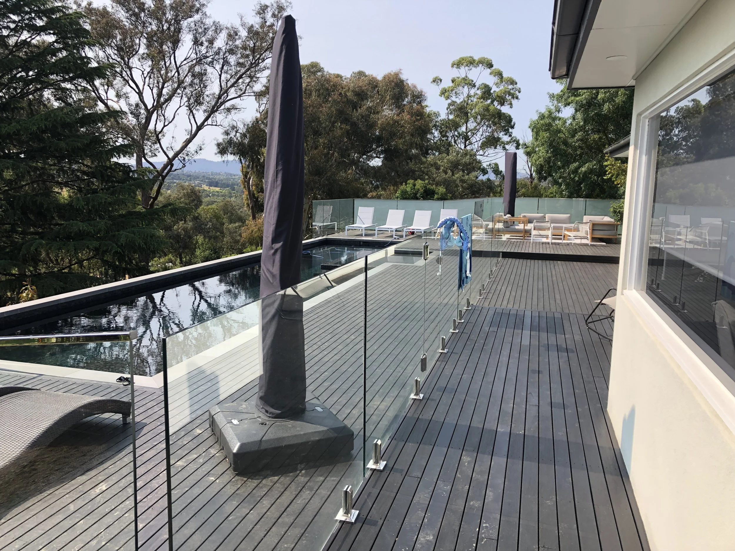 Modern rooftop deck with pool, lounge chairs, umbrellas, surrounded by glass railing and trees in the background.