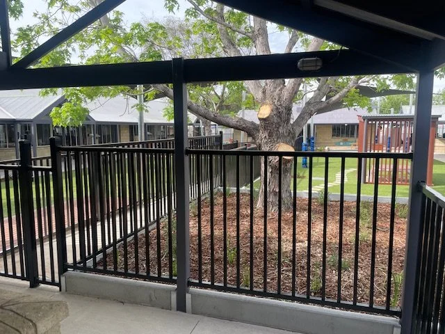 View from a porch showing a fenced backyard with a large tree and a house in the background.